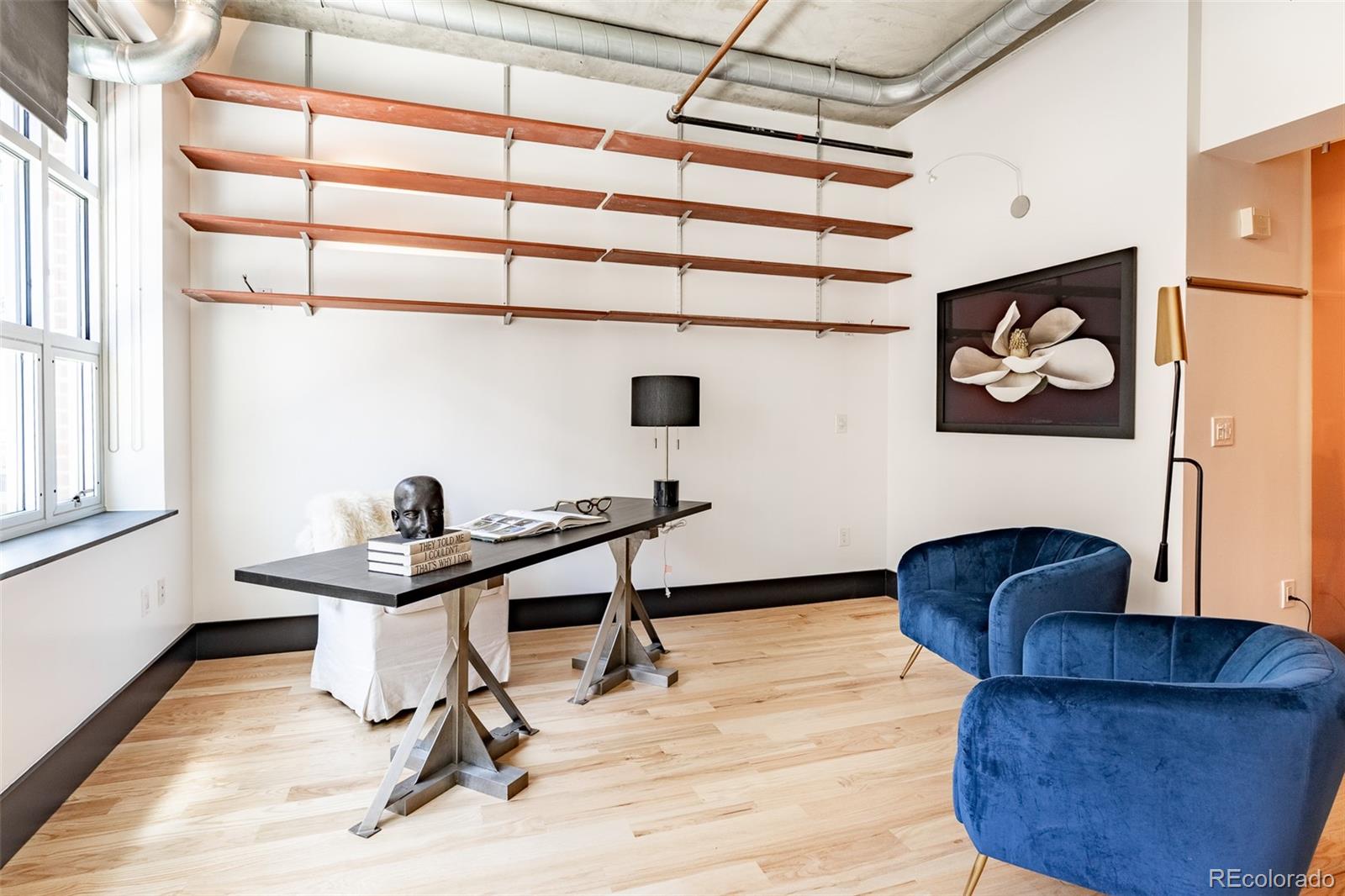 Loft-style home office with long desk, wall-mounted wood shelves, and blue velvet chairs.