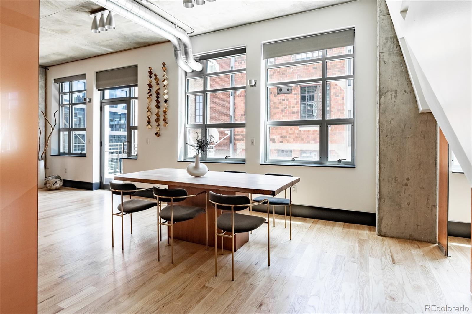 Loft dining area with a wood table, large windows, pale floors, and exposed concrete details.