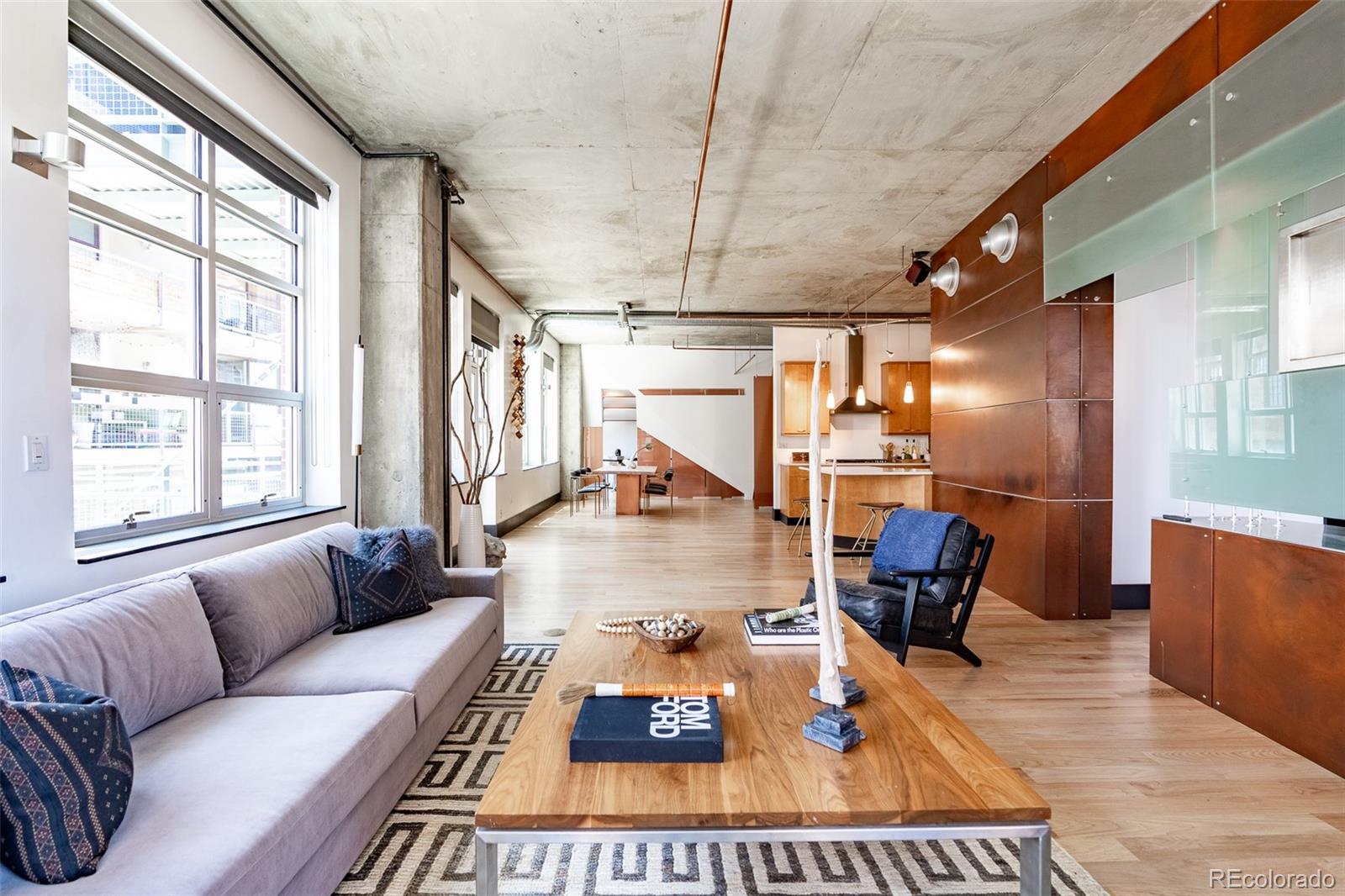 Loft living room with large windows, exposed concrete, gray sofa, and open kitchen.
