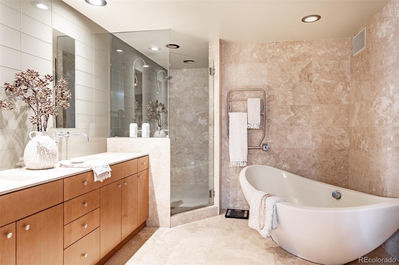 Bathroom with a glass shower, freestanding tub, and long wood vanity against beige stone tile.