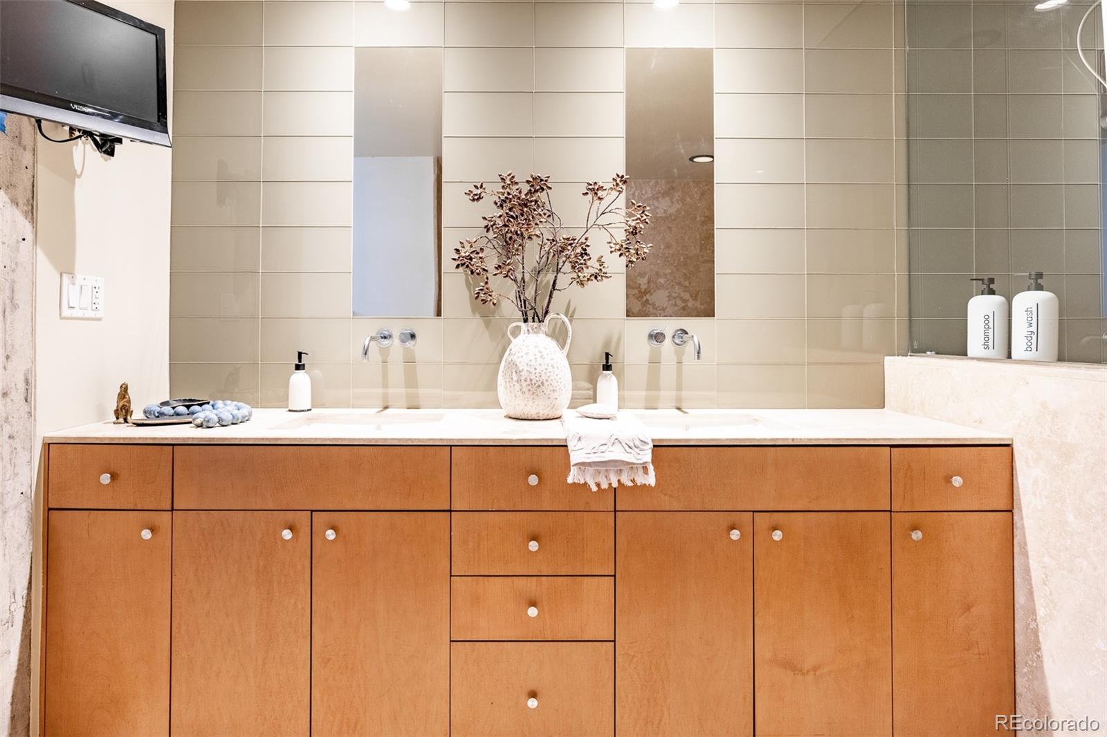 Bathroom vanity with two sinks, warm wood cabinetry, beige tile and a glass shower enclosure.