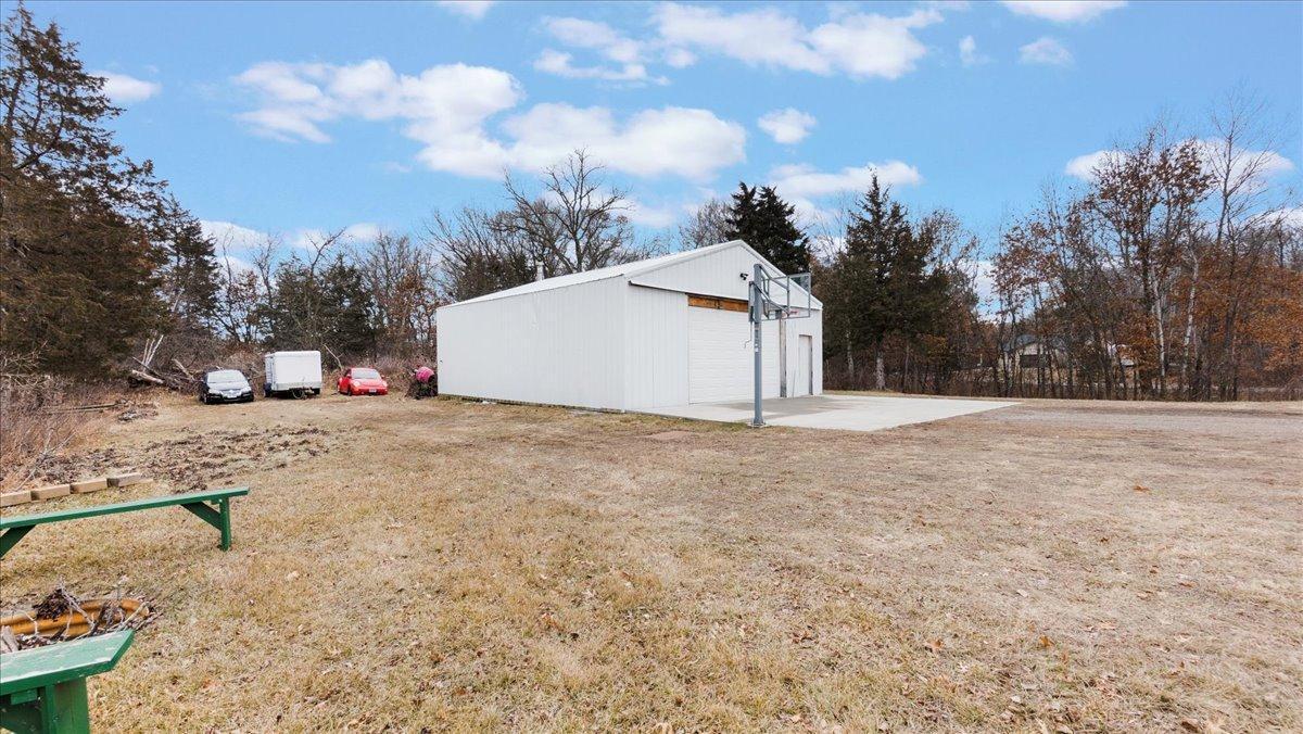 White metal outbuilding with a basketball hoop beside a broad paved area and parked vehicles.