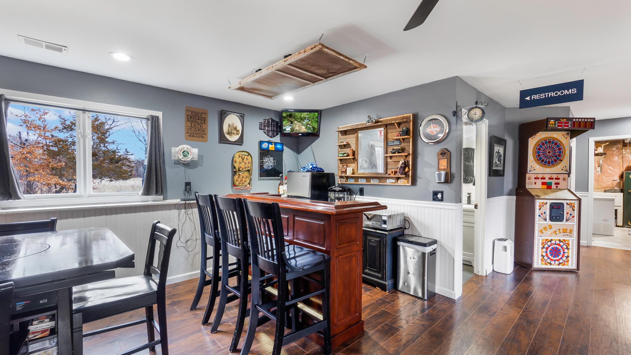 Recreation room with a wood bar, dart machine, and window overlooking a lightly snow-covered yard.