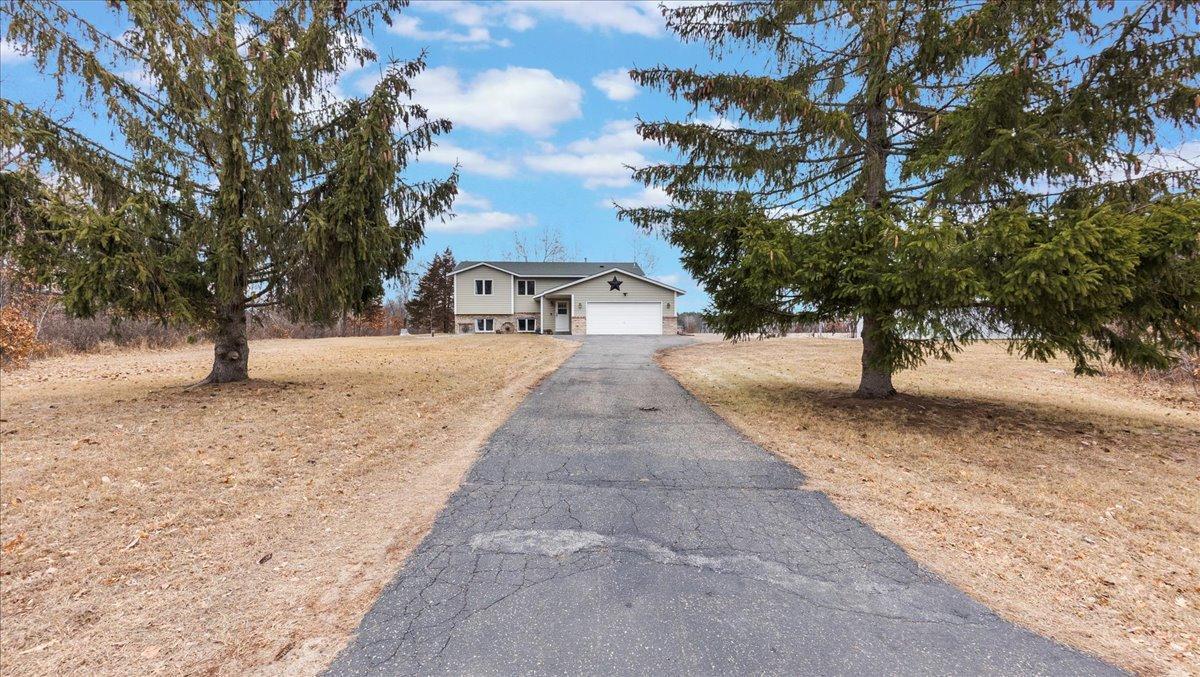 Two-story home with an attached garage at the end of a long driveway, framed by large evergreen trees.
