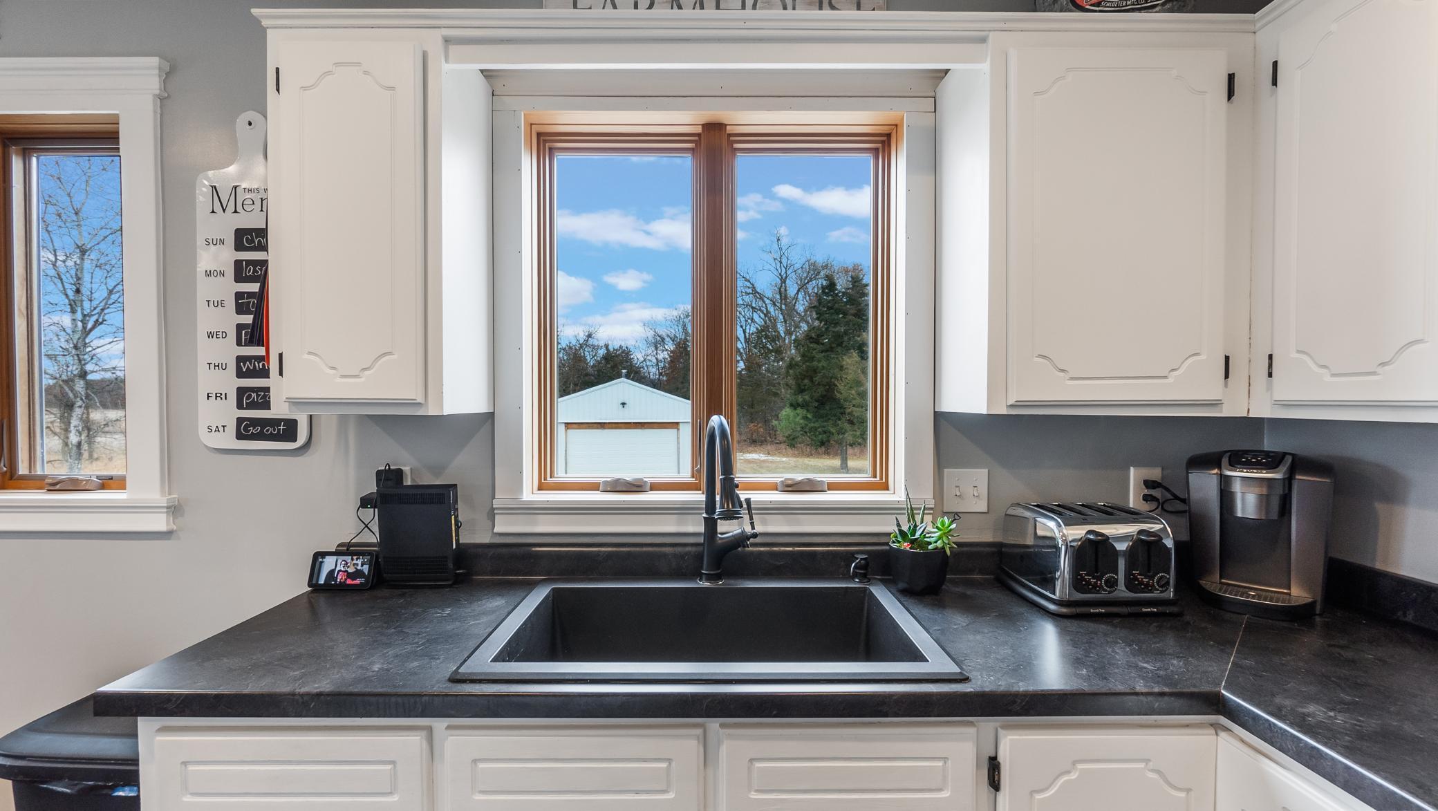 Kitchen with a large black sink beneath wood-trimmed windows and white cabinets.