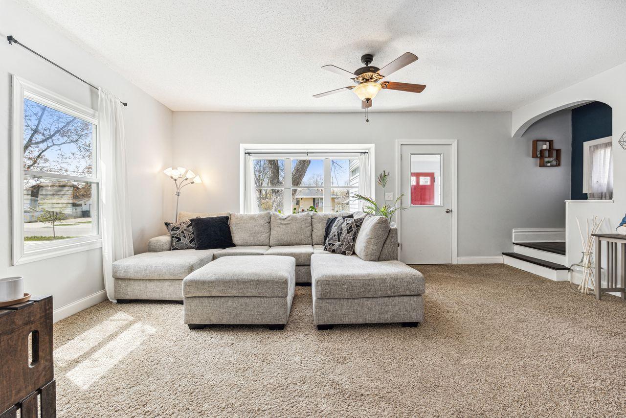 Living room with a light-gray sectional, beige carpet, two windows, and a ceiling fan.