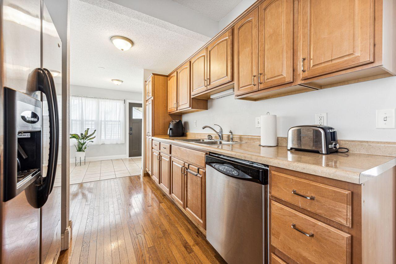 Kitchen with honey-toned wood cabinets, stainless steel appliances, and hardwood floors leading to a tiled entry.