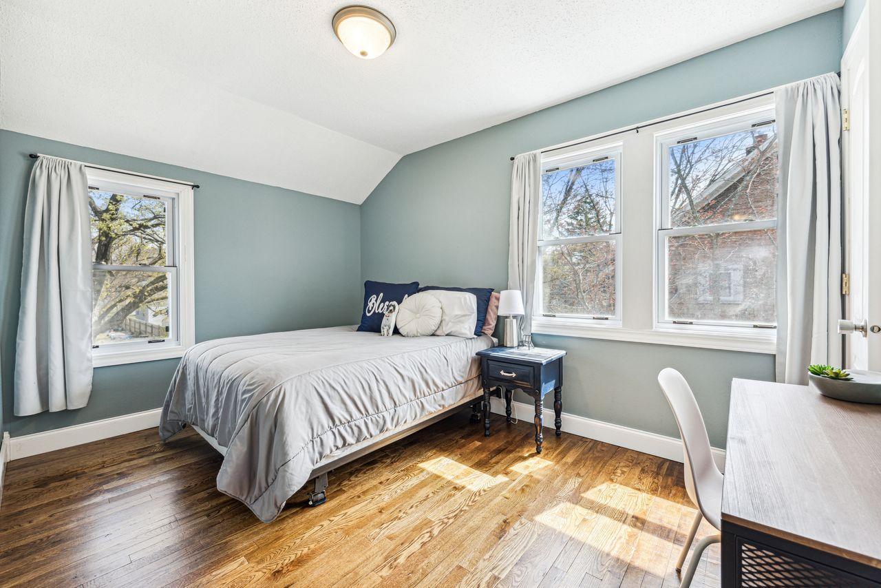 Bedroom with gray-bedded bed, blue-green walls, three windows, and sunlit hardwood floors.