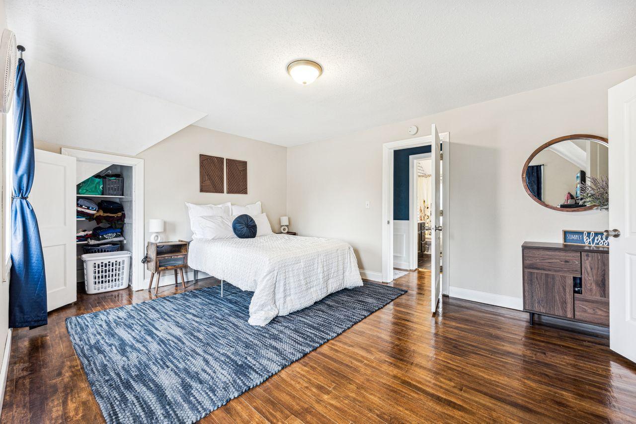 Bedroom with a white bed, blue patterned rug, open closet, and dark wood floors.