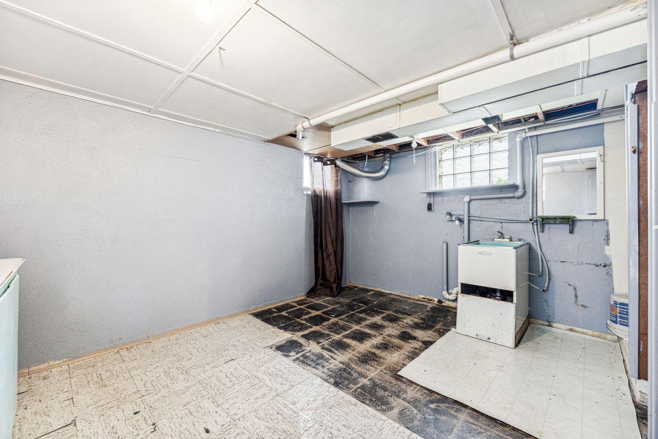 Basement laundry room with a utility sink, exposed ductwork, glass-block window, and worn flooring.