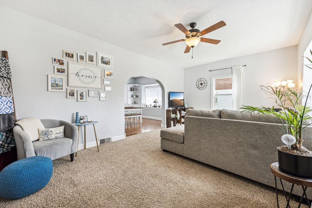 Furnished living room with a gray sectional, beige carpet, framed photos, and an arched dining-area opening.