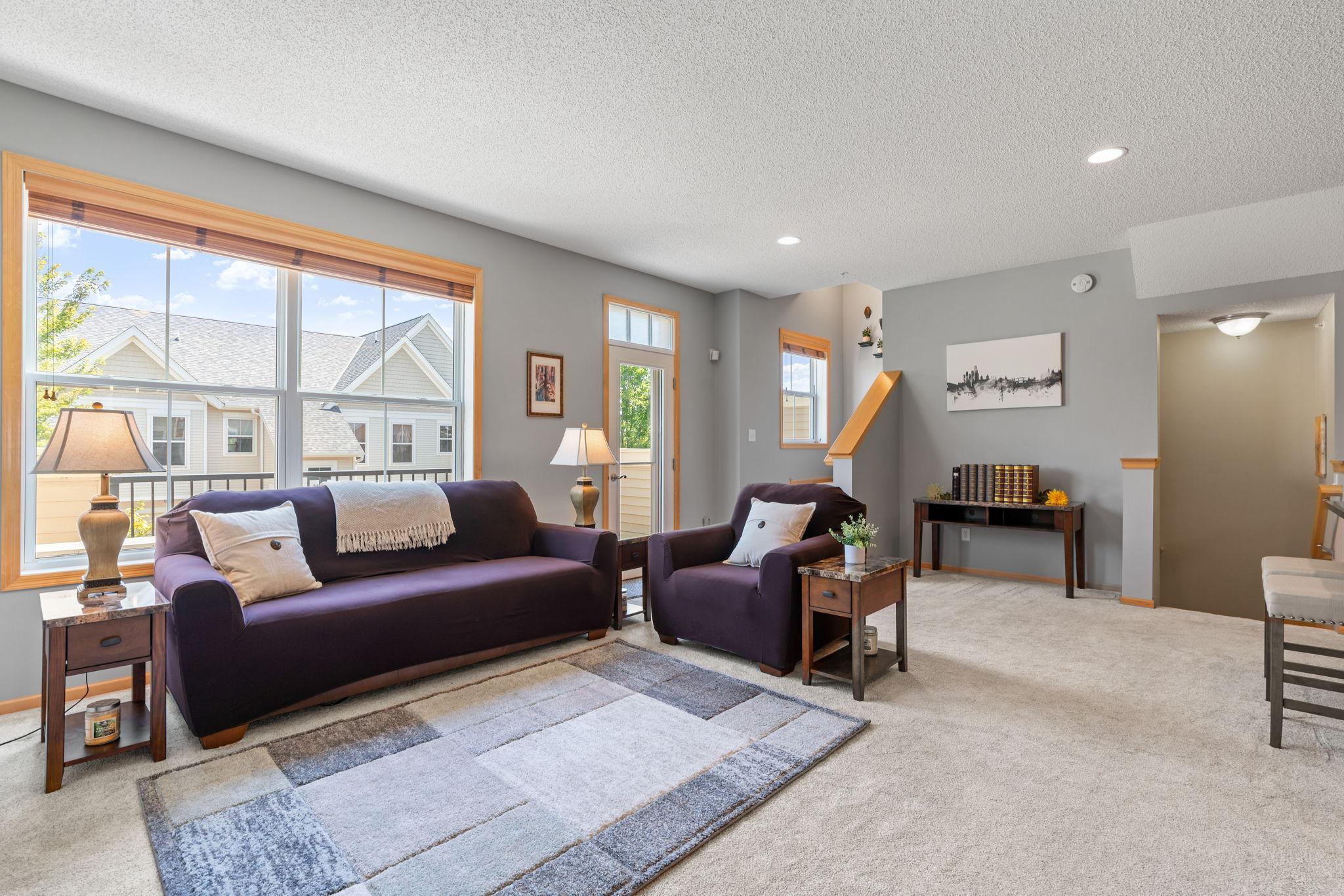Furnished living room with a purple sofa, matching armchair, and large windows overlooking neighboring homes.
