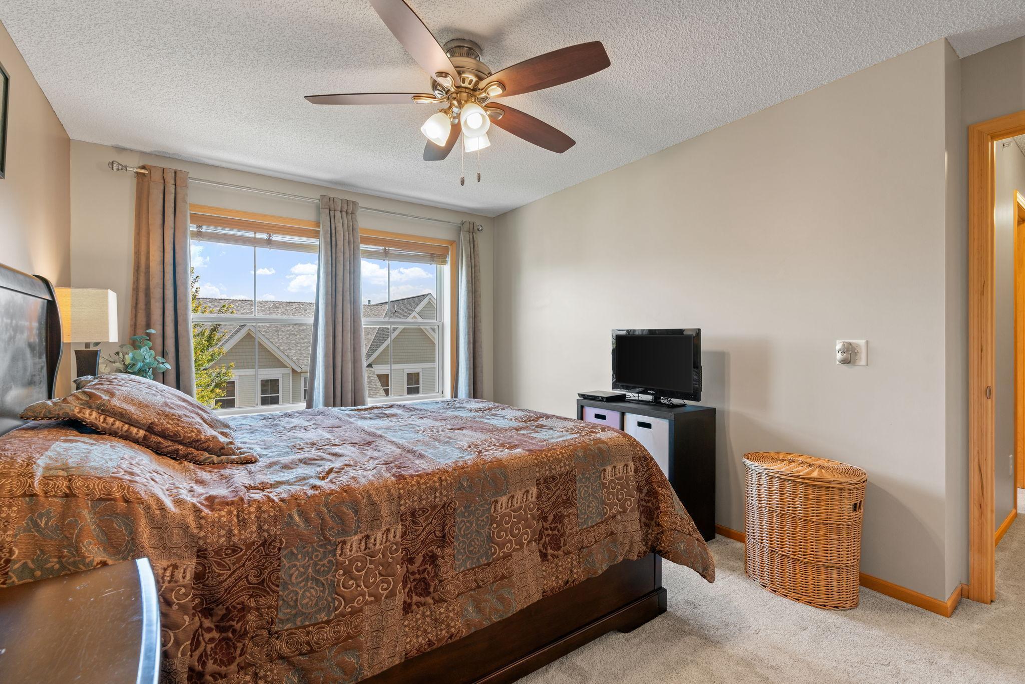 Bedroom with a patterned bedspread, ceiling fan, and window overlooking neighboring homes.