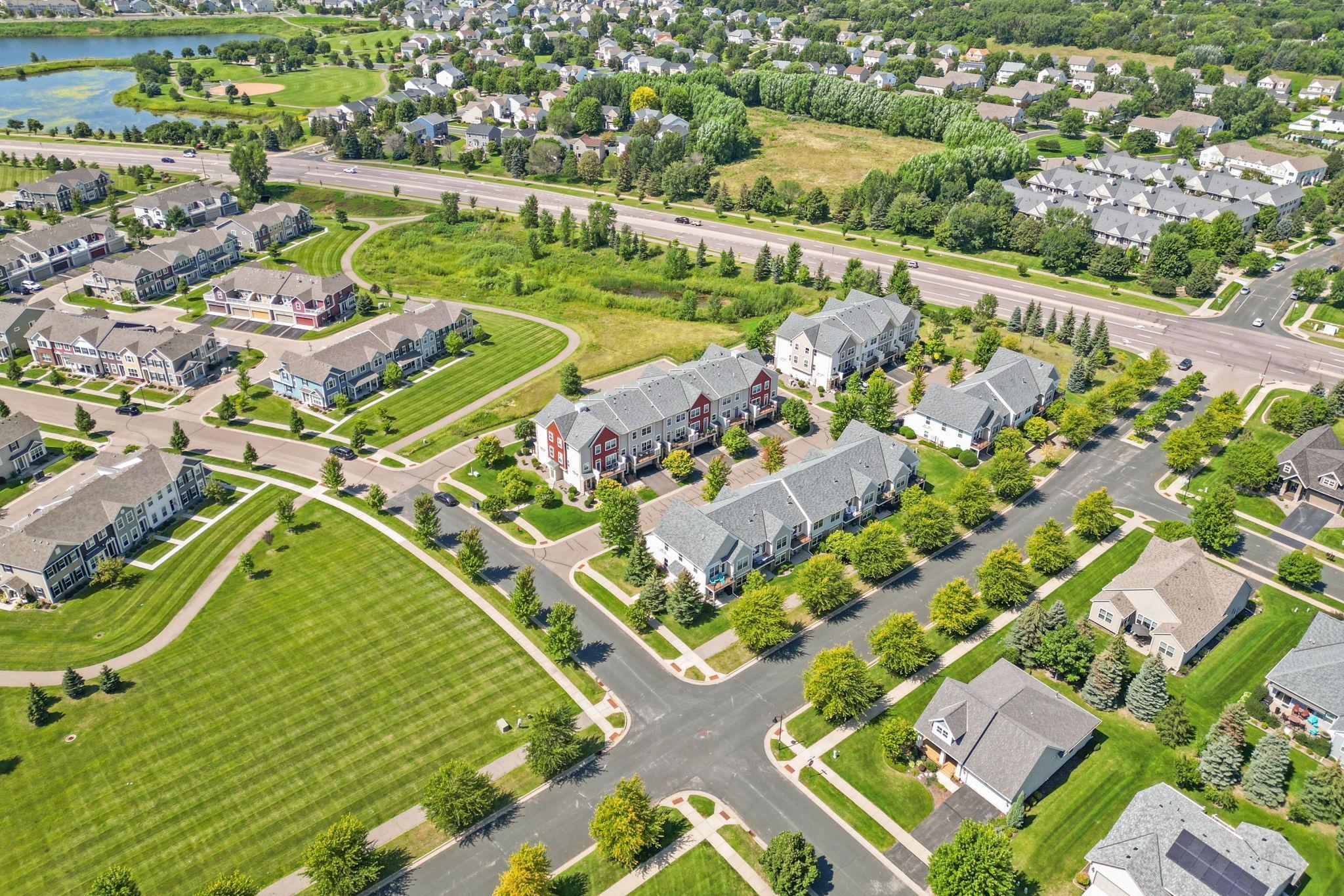 Aerial view of townhomes and landscaped lawns, with a lake and major road in the distance.