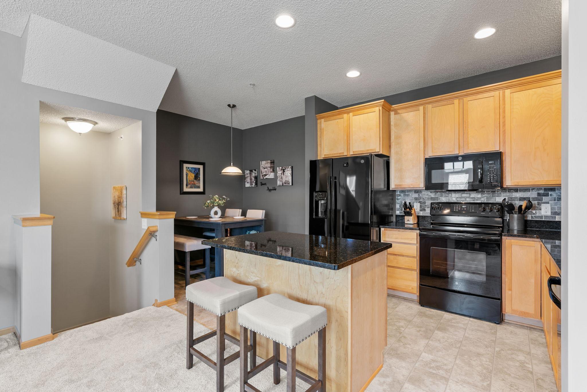Condo kitchen with a dark stone island, light wood cabinets, and black appliances.