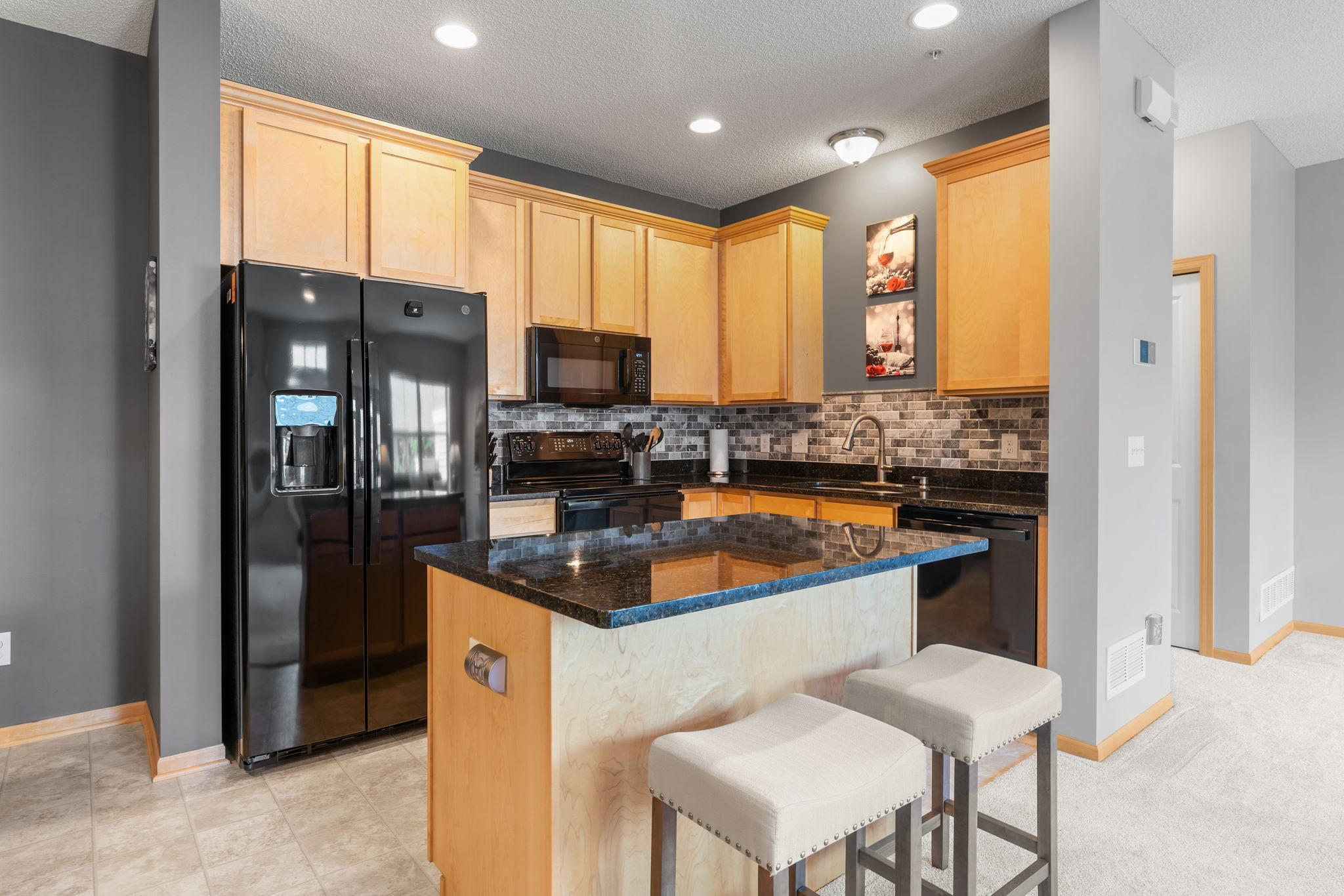 Kitchen with light wood cabinets, black appliances, and a stone island with two upholstered stools.