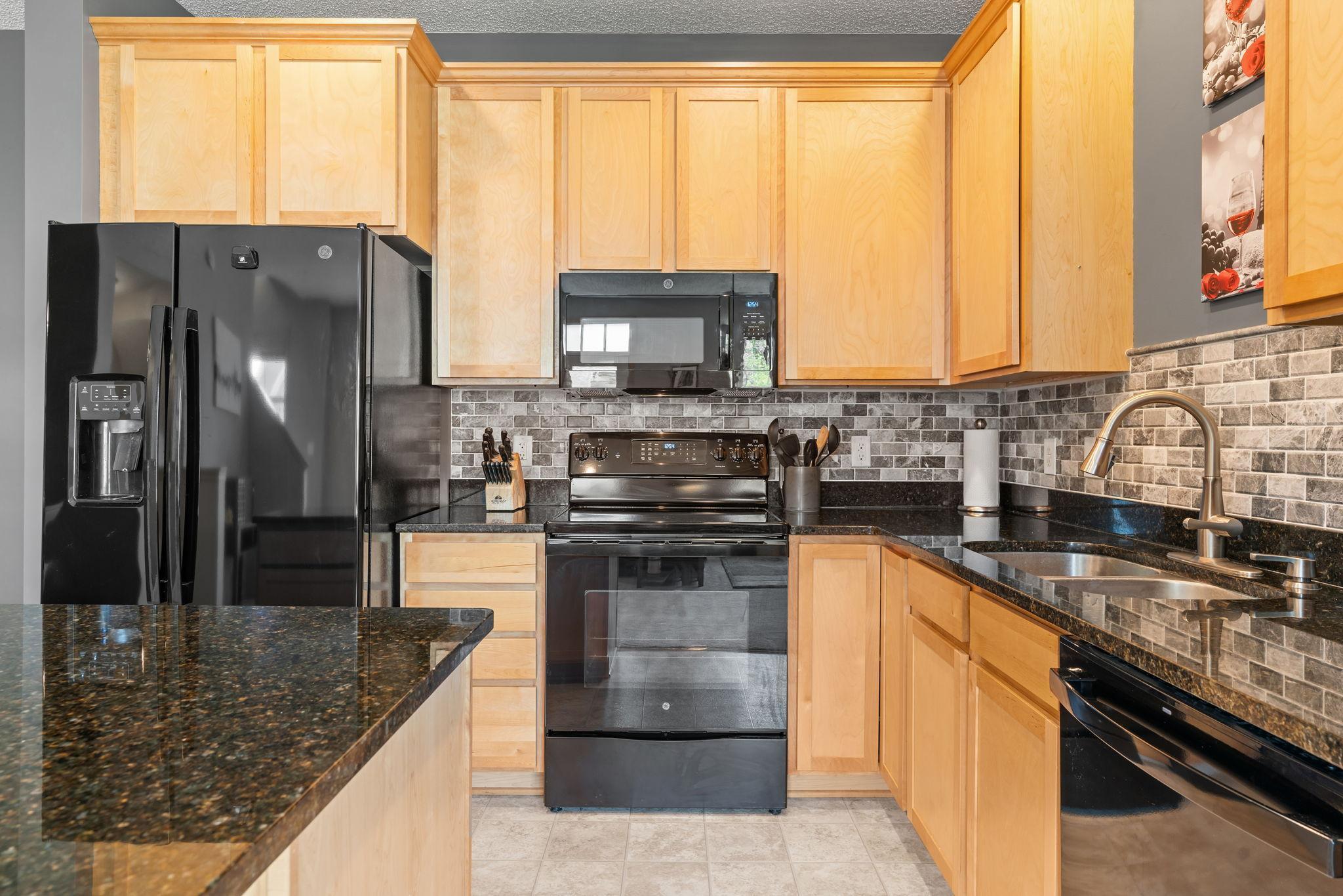 Kitchen with light wood cabinets, dark granite counters, gray tile backsplash, and black appliances.