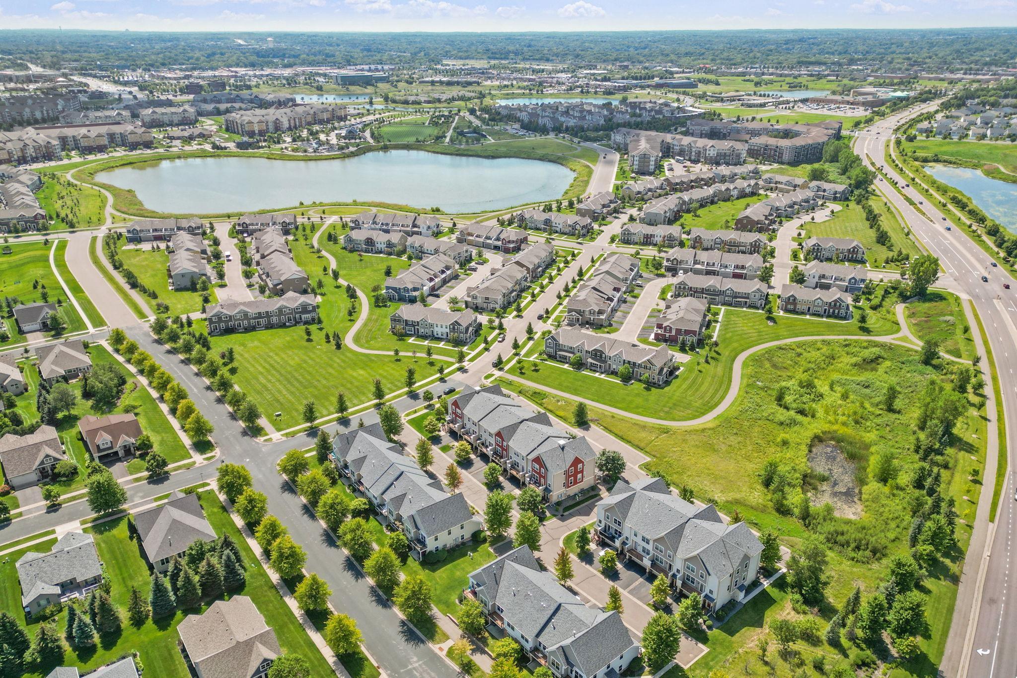 Aerial view of a townhouse community with a large pond, landscaped paths, and surrounding roads.