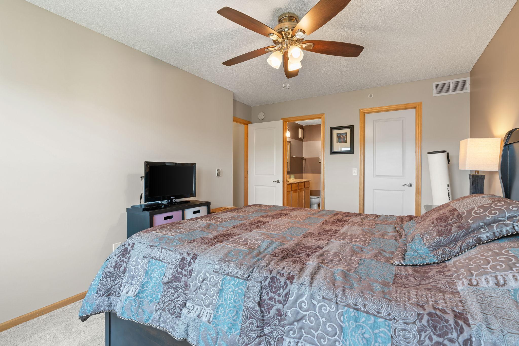 Bedroom with a patterned quilt, ceiling fan, television, and open doorway to an adjoining bathroom.