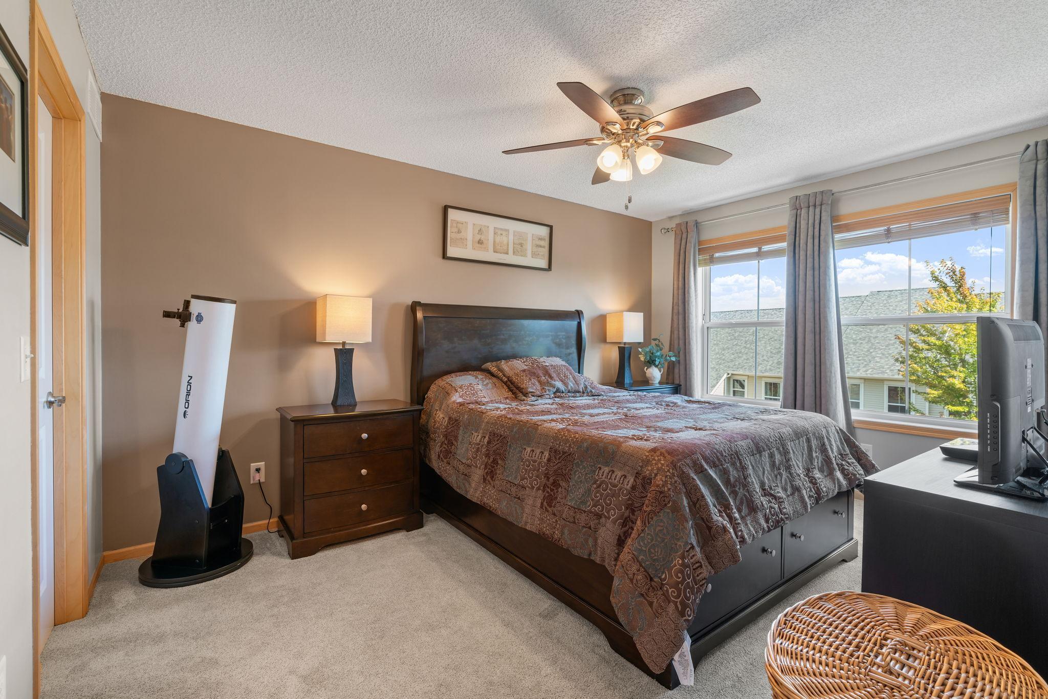 Bedroom with a dark wood bed, patterned bedding, ceiling fan, and large curtained window.