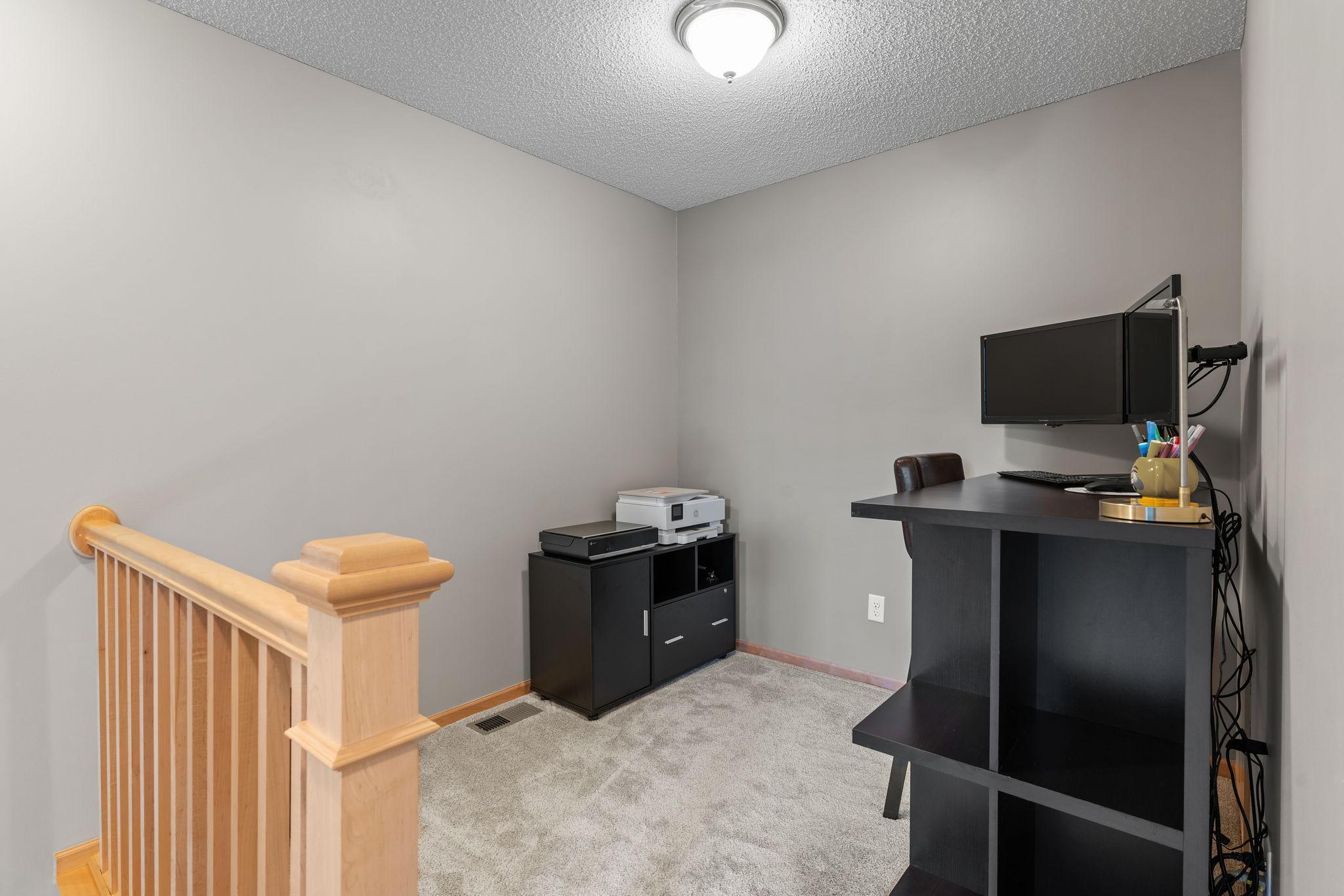 Furnished home office with a black desk and monitors beside a wooden stair railing.