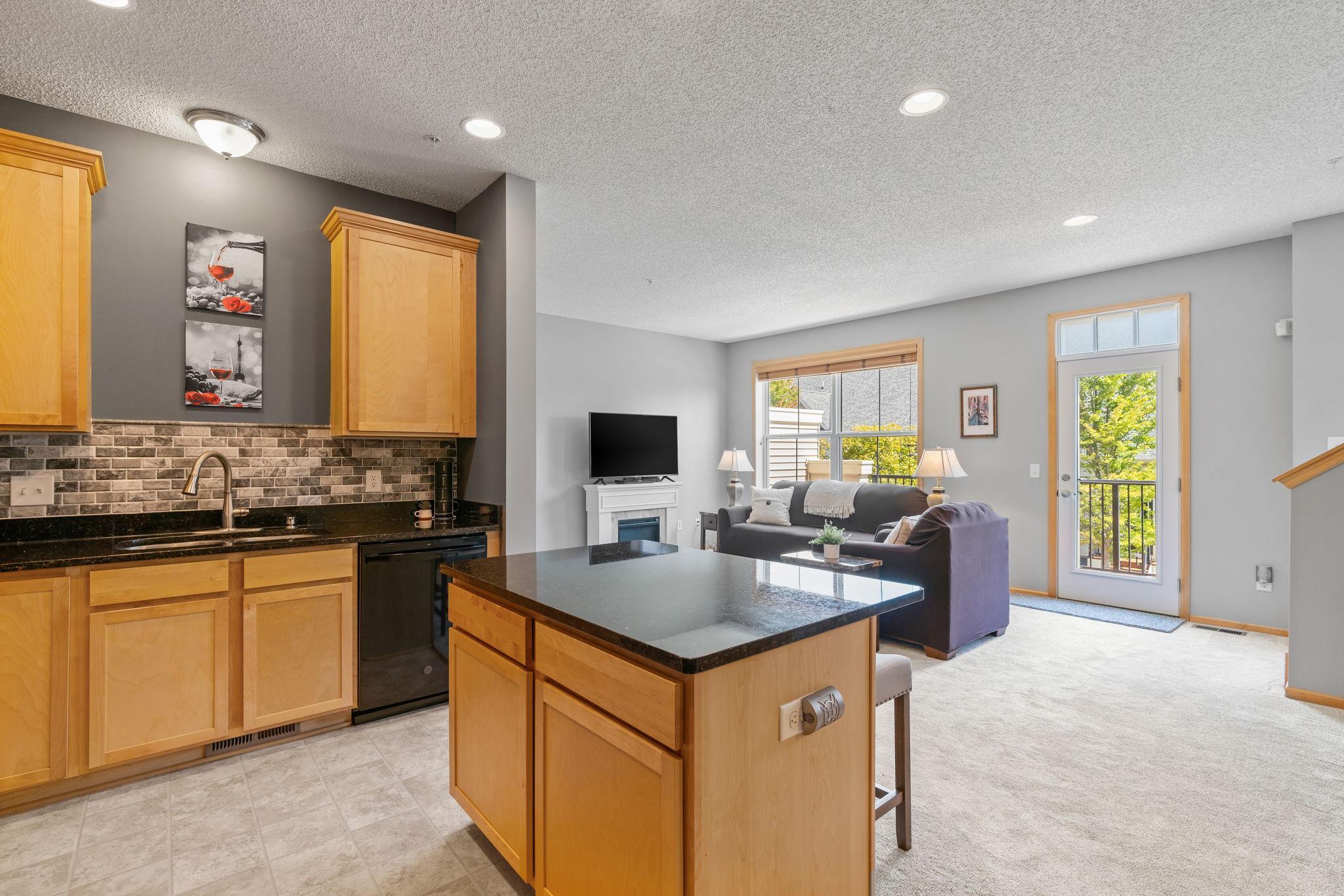 Open kitchen and living room with a dark stone island and wood cabinetry beside a bright balcony door.