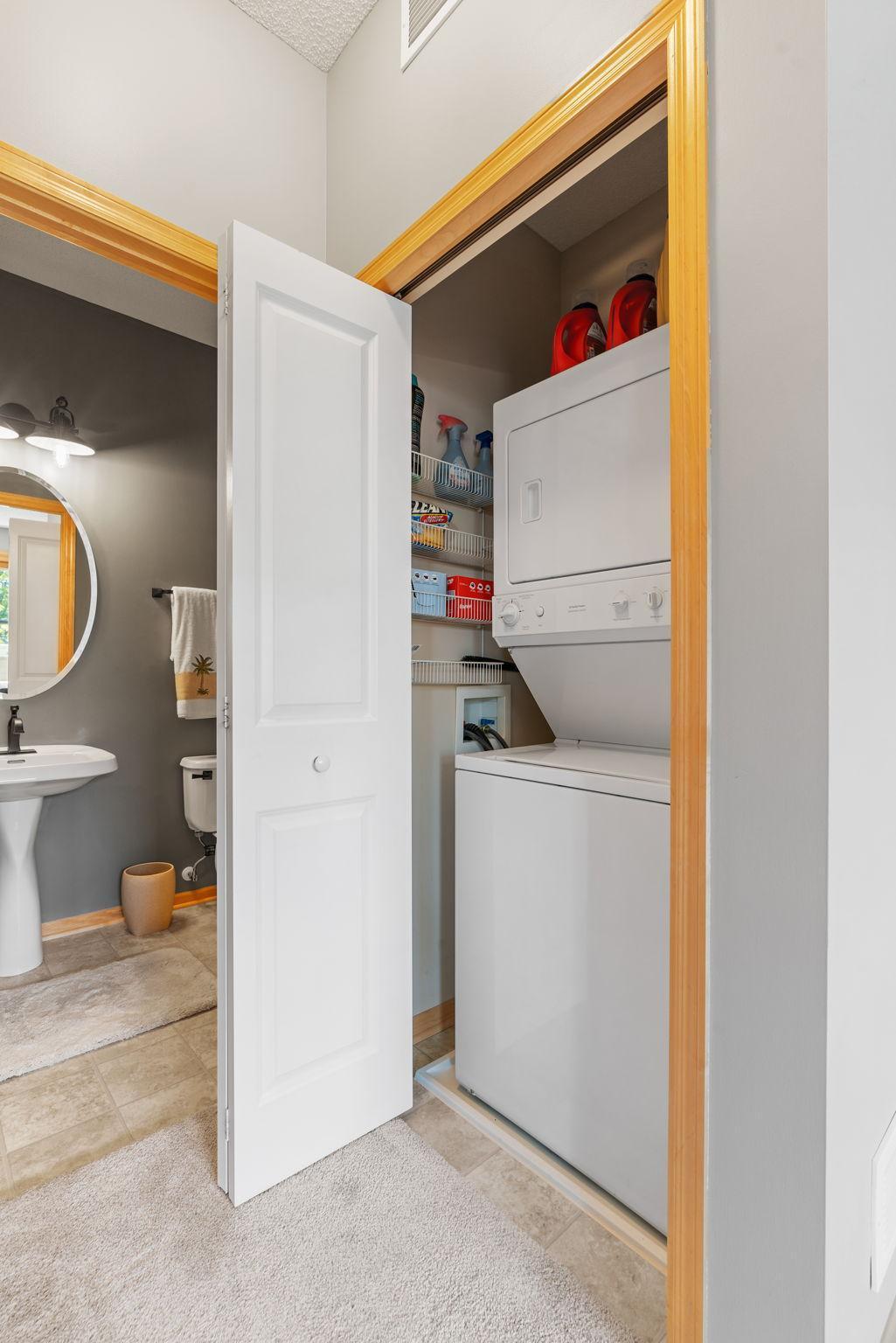 Laundry closet with stacked washer and dryer and open storage shelves beside a small bathroom.