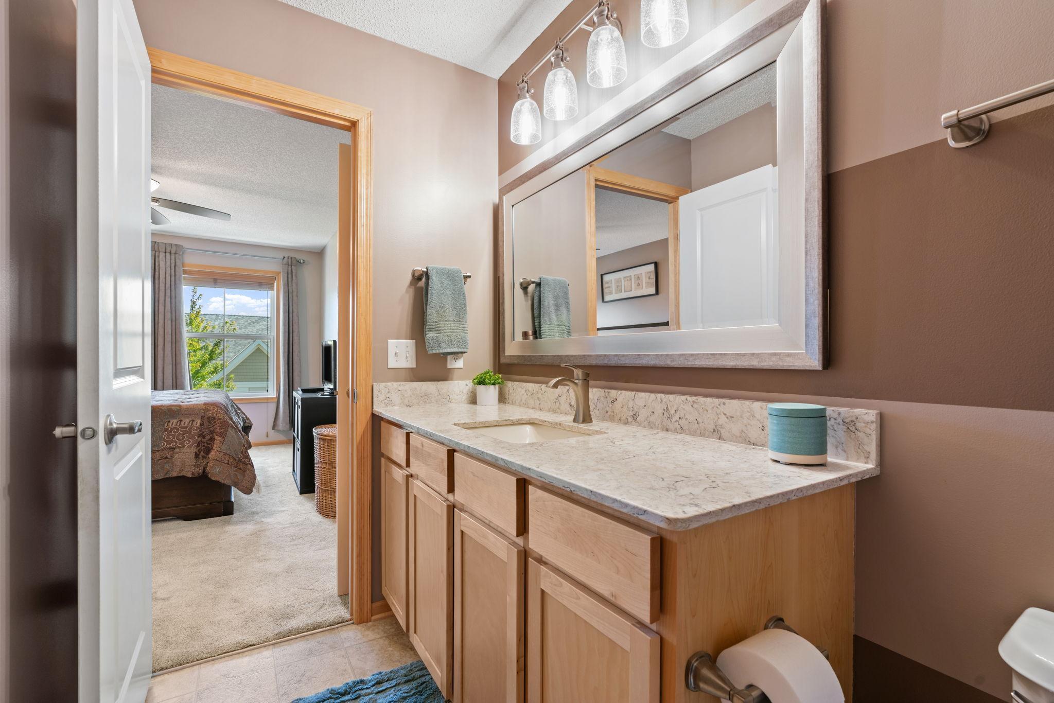 Bathroom with a long wood vanity and stone counter, looking through an open doorway into a sunlit bedroom.
