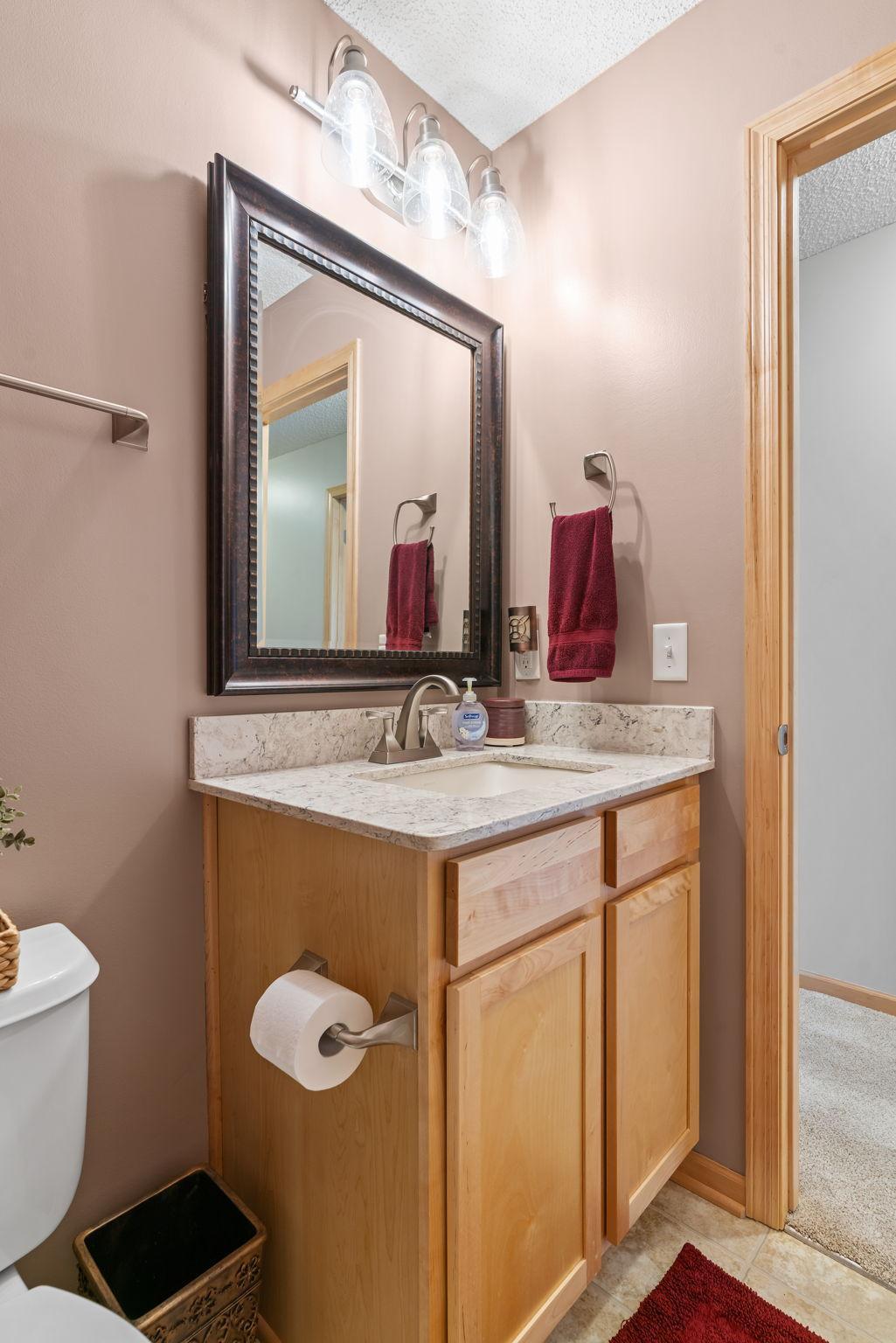 Bathroom vanity with light wood cabinetry, stone countertop, framed mirror, and red towels.