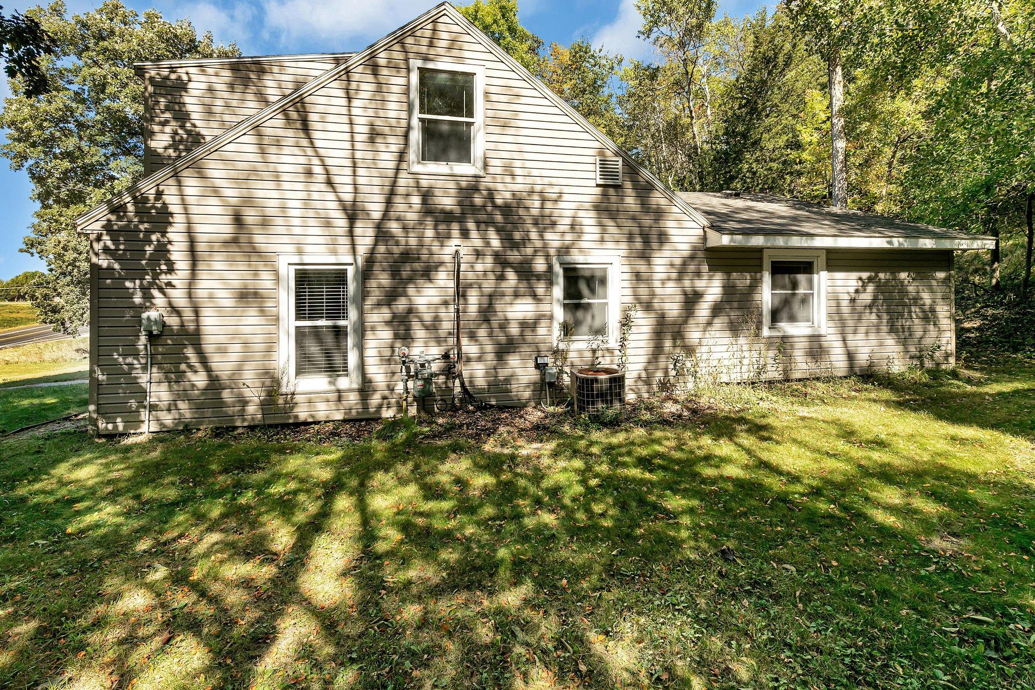 Home’s beige-sided exterior with a steep gable, outdoor utility equipment, and a tree-shaded lawn.