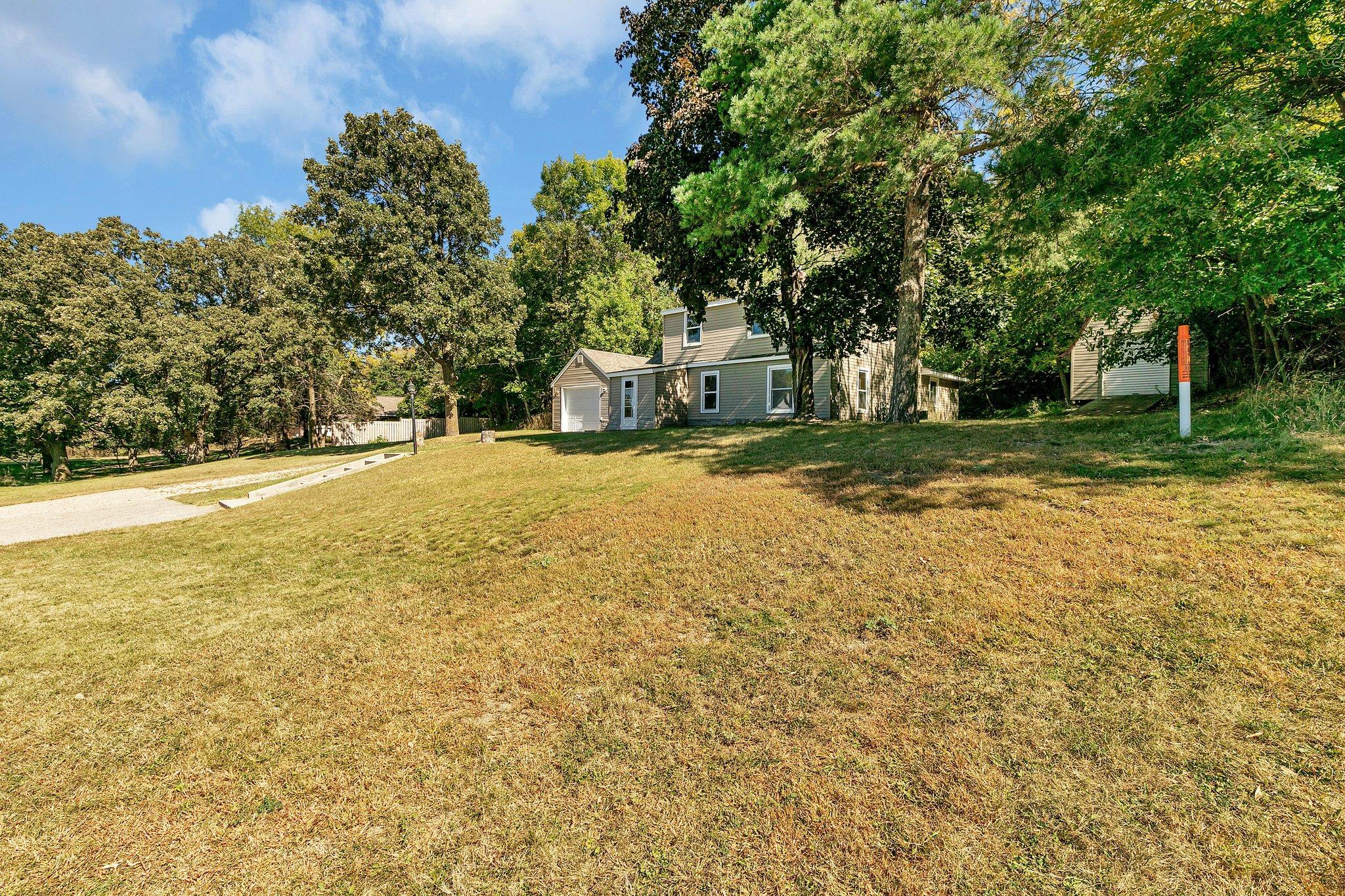 Beige two-story house with attached garage among mature trees, above a broad sloping lawn.