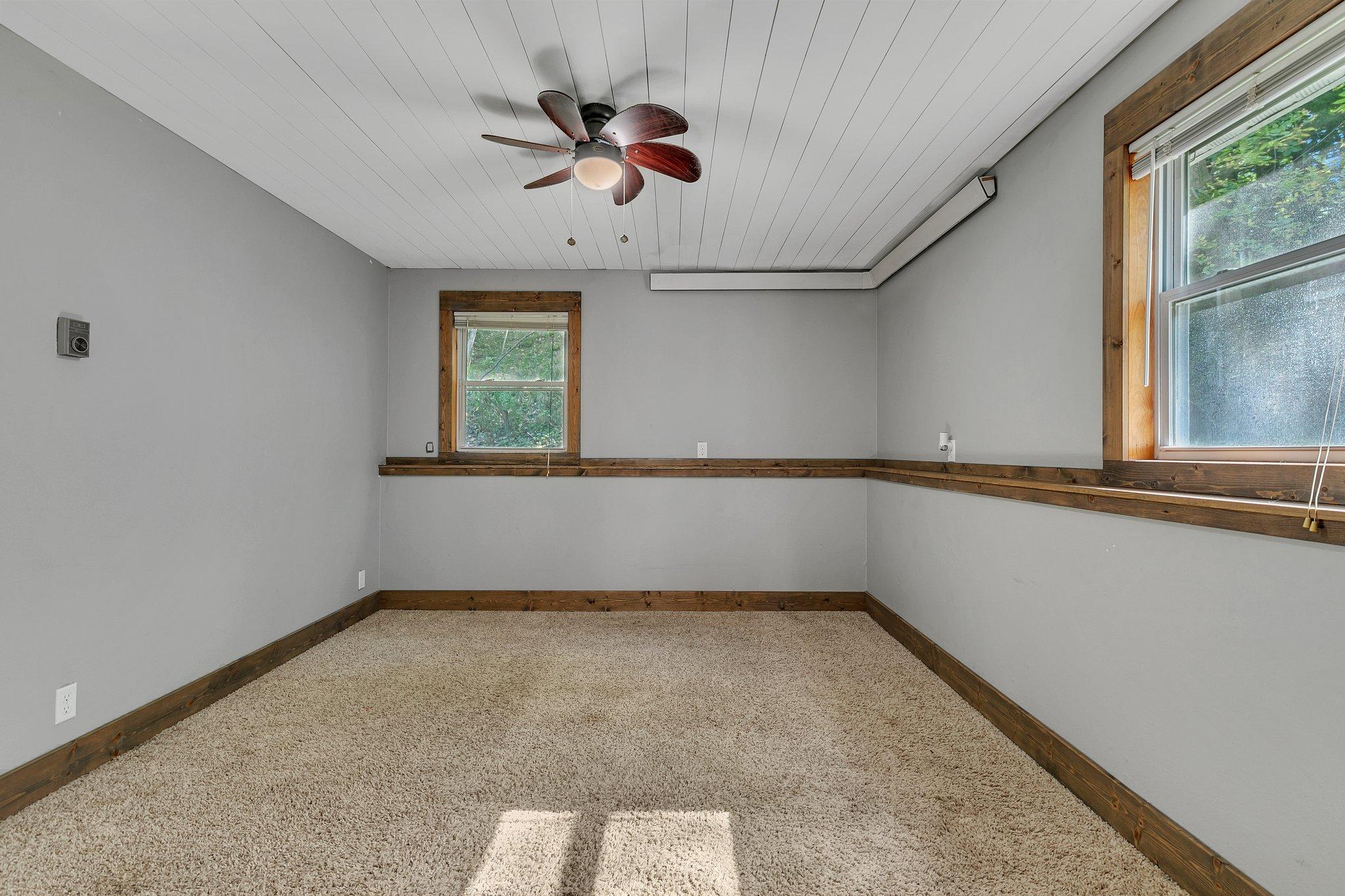Empty carpeted room with dark wood trim, two windows, and a ceiling fan.