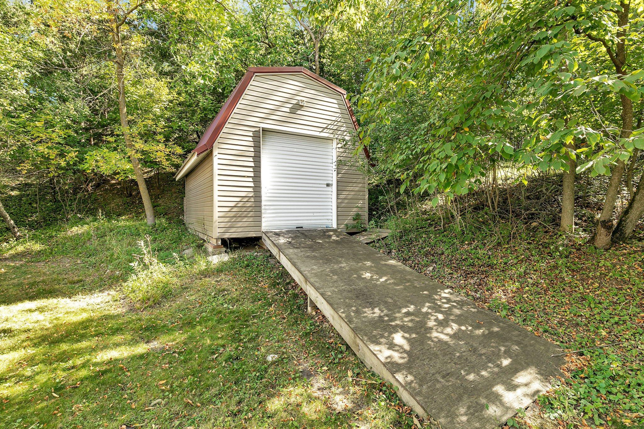 Small beige shed with a white roll-up door and long wooden ramp in a wooded yard.