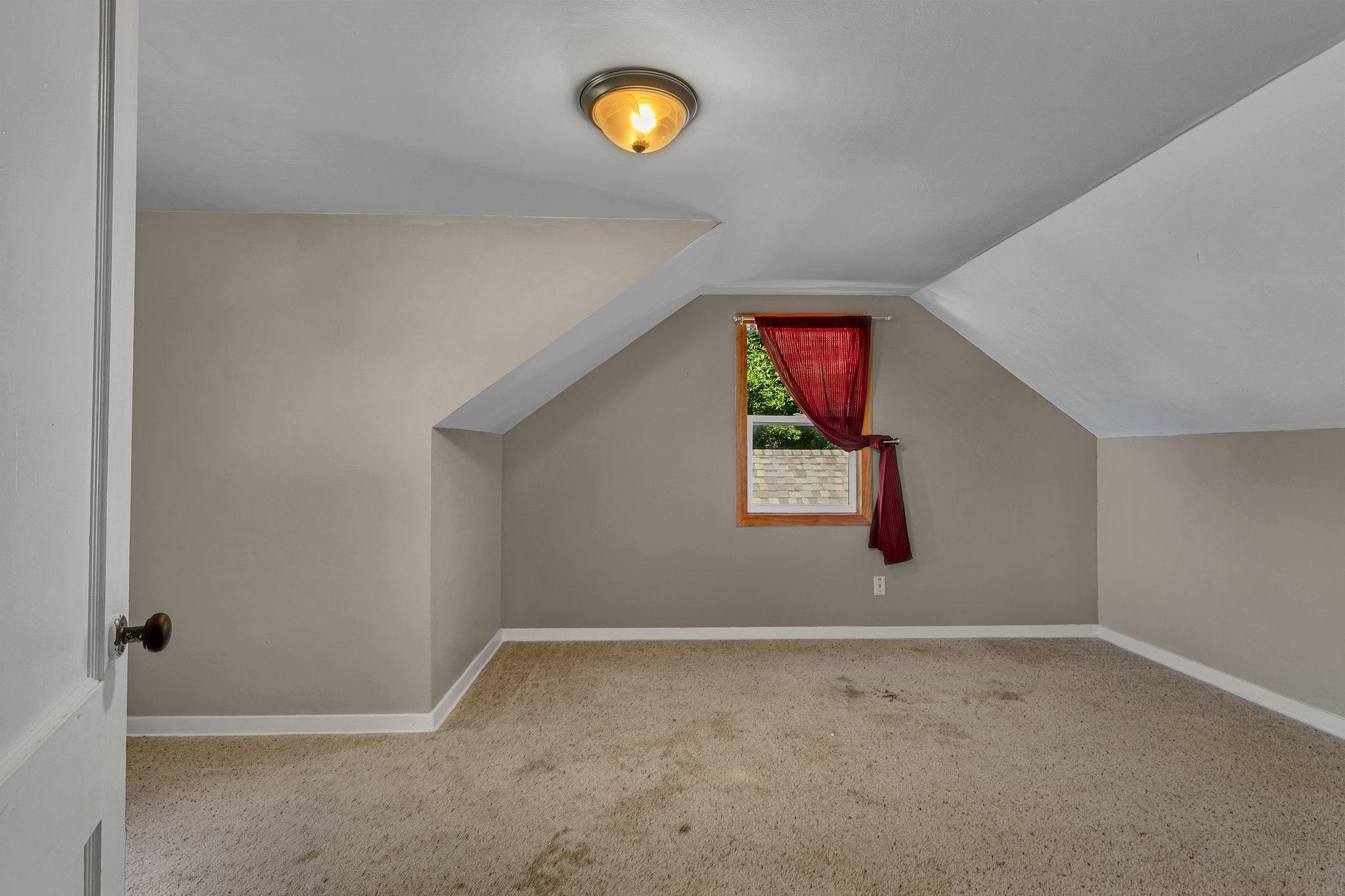 Empty carpeted upper-level room with sloped ceilings and a small window framed by a red curtain.