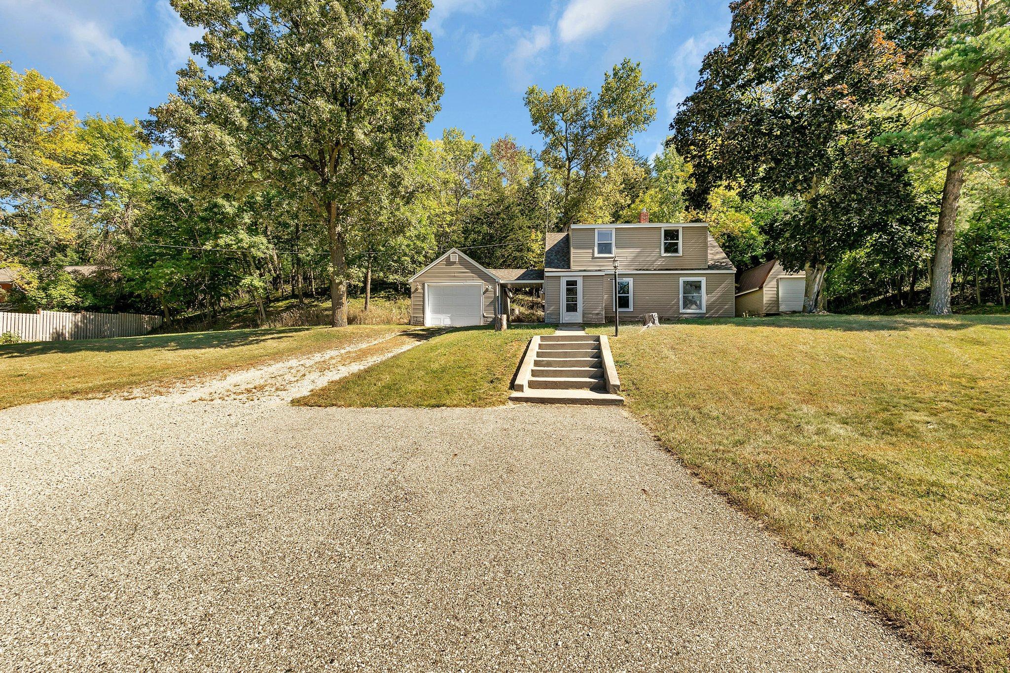 Two-story beige house with an attached garage, gravel driveway, and mature trees surrounding the yard.