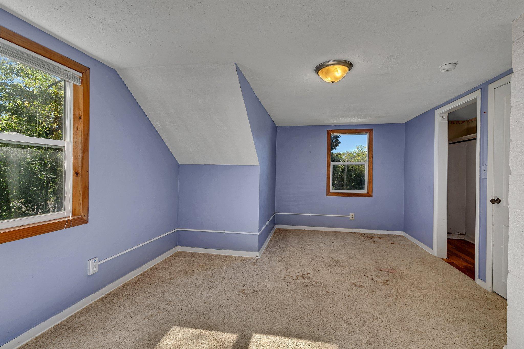 Empty lavender bedroom with a sloped ceiling, beige carpet, and wood-trimmed windows.