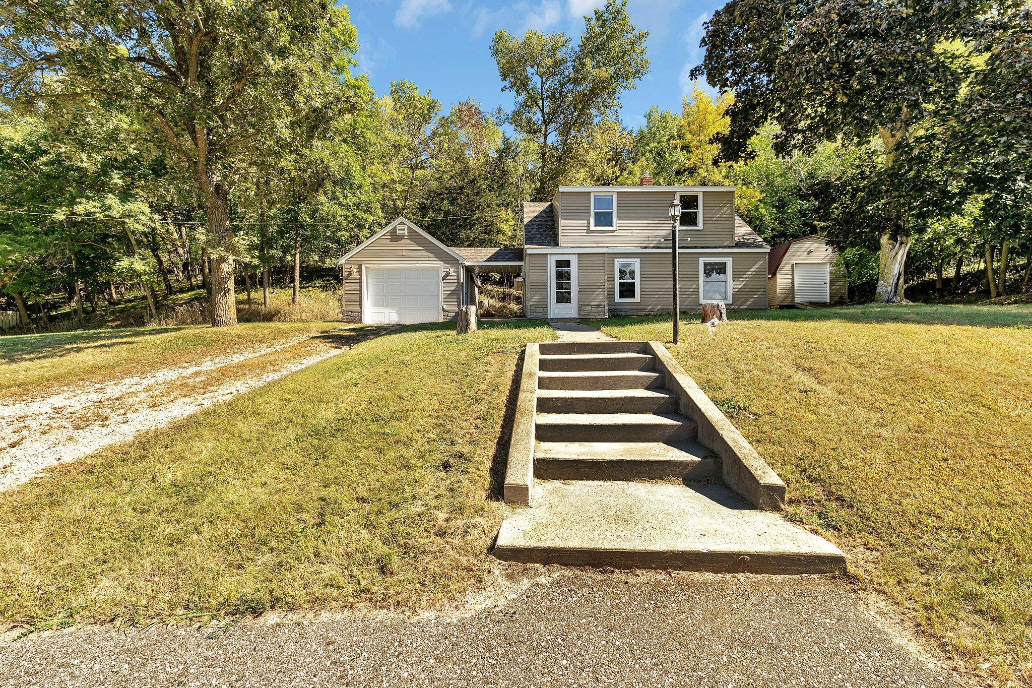 Tan two-story house with attached garage, detached shed, and concrete steps in a wooded yard.