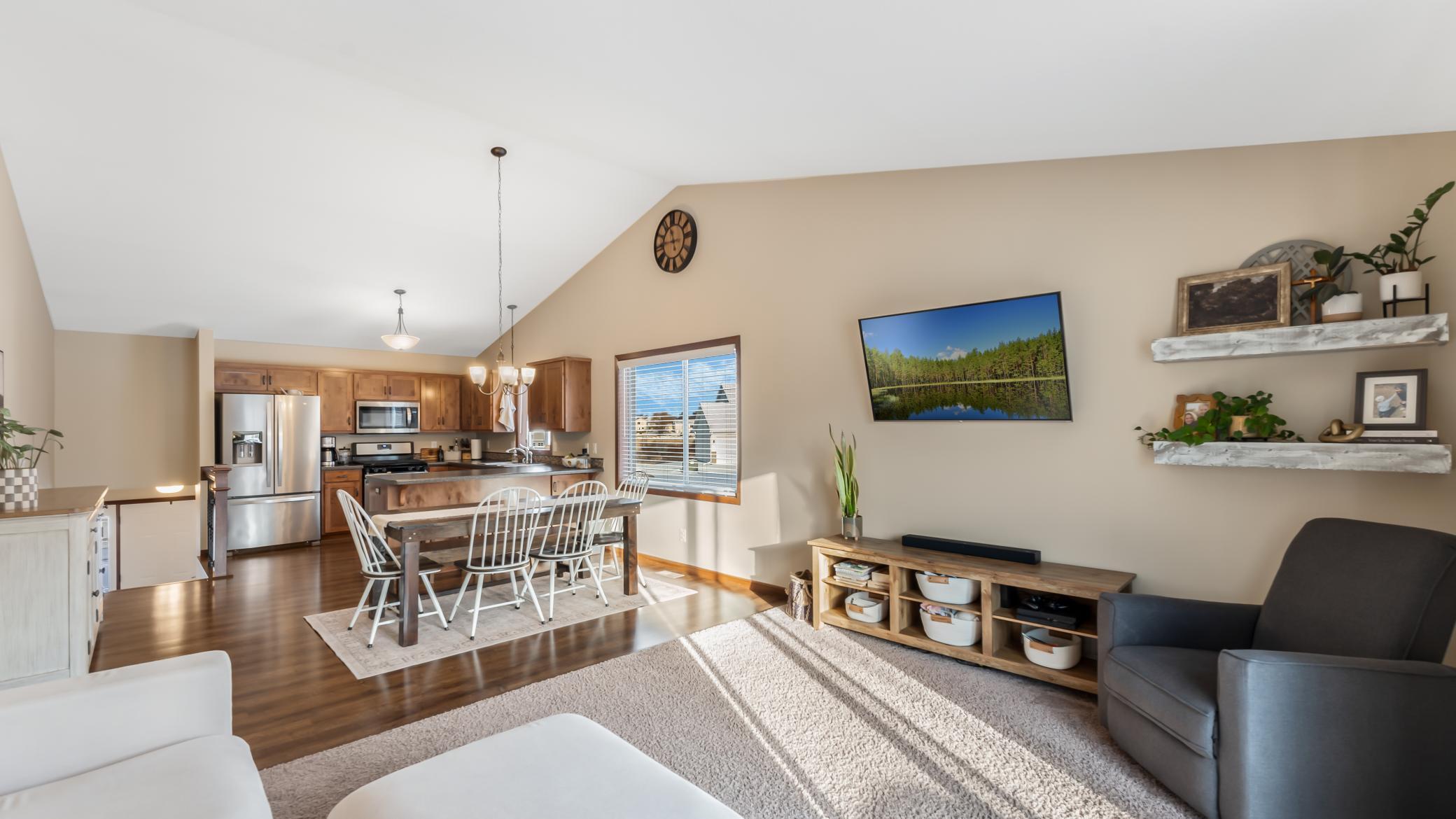 Open-plan living room and kitchen with wood cabinets, a dining table, and sunlight across the floor.