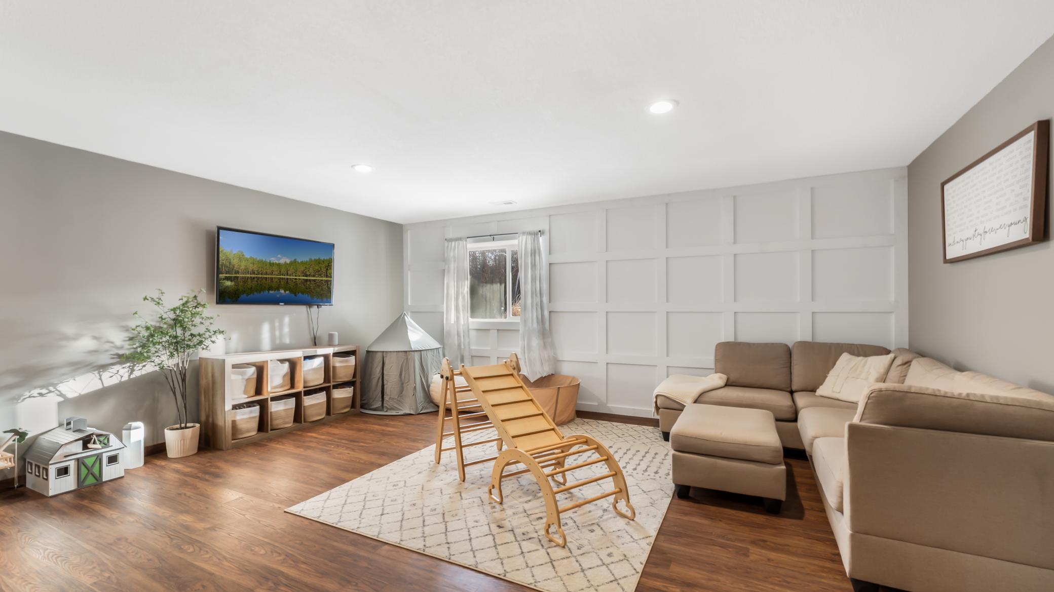 Furnished living and playroom with a white paneled wall, beige sectional, and wooden rocking toy.