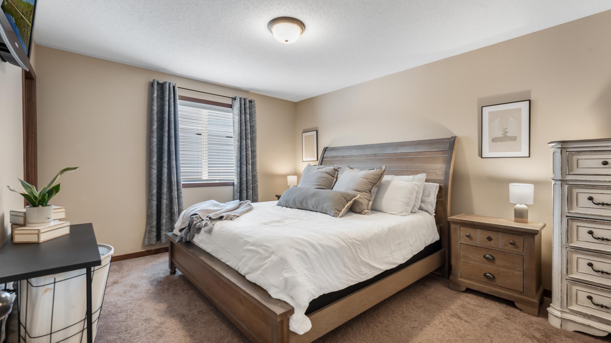 Staged bedroom with a wooden bed, layered gray and white pillows, and gray curtains framing the window.