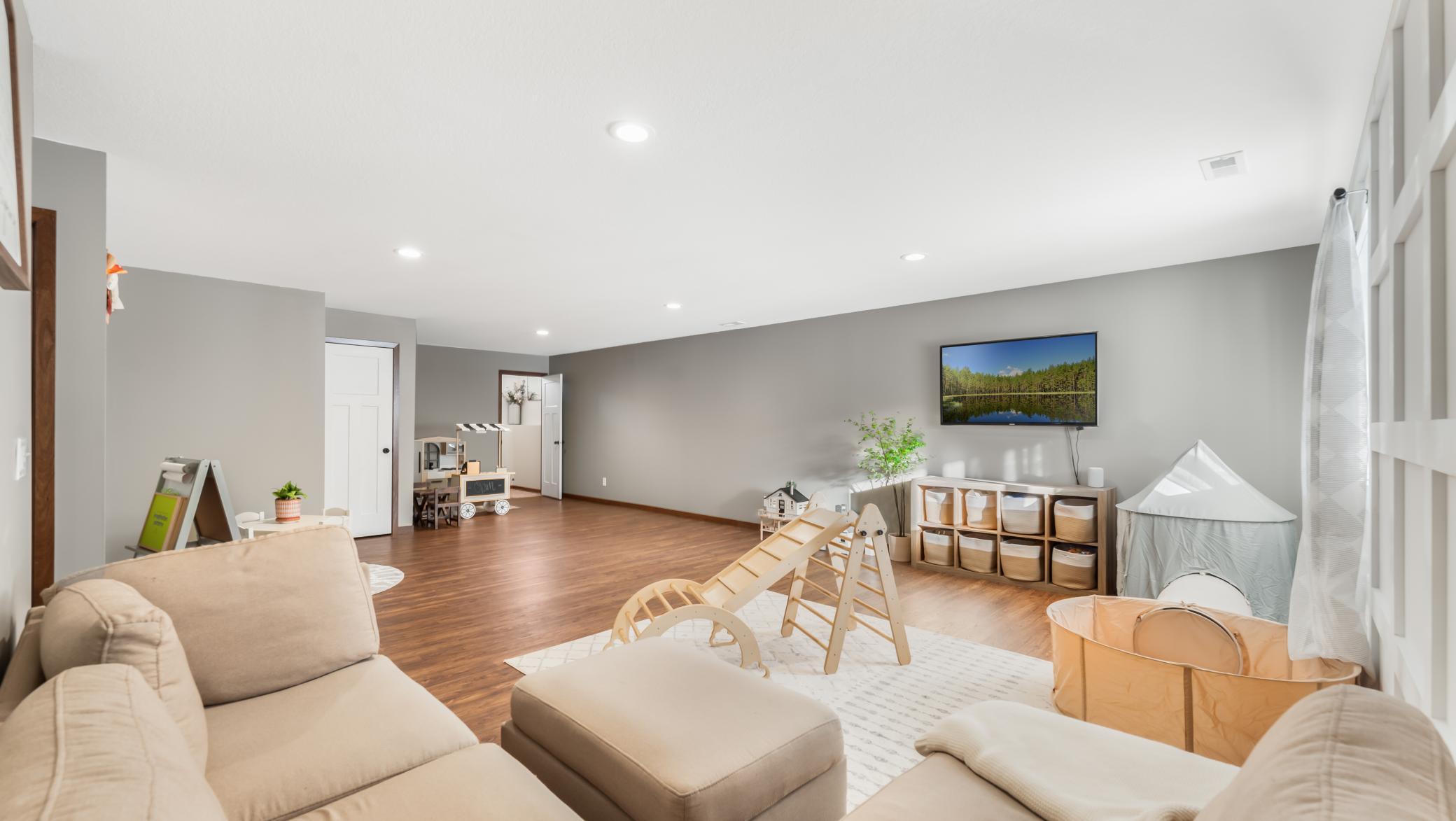 Furnished playroom with a beige sectional, children’s climbing equipment, toy storage, and wall-mounted TV.