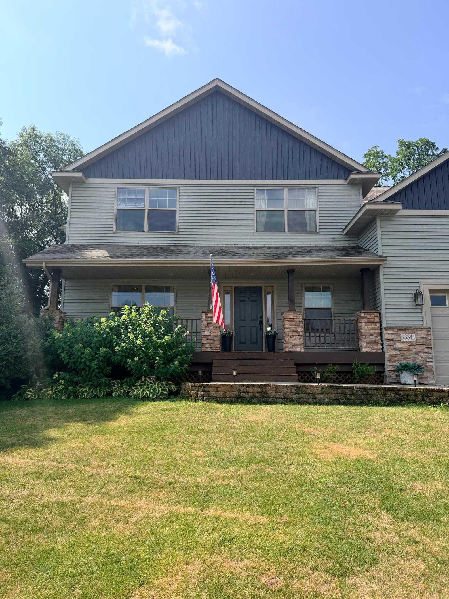 Two-story gray house with a blue gable, stone porch pillars, American flag, and broad lawn.