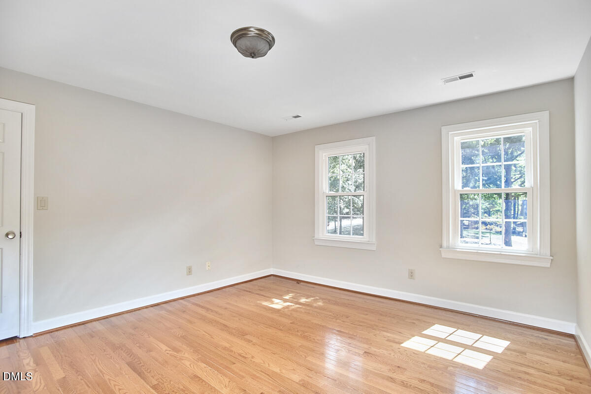 Vacant room with hardwood floors, neutral walls, and two windows overlooking trees.