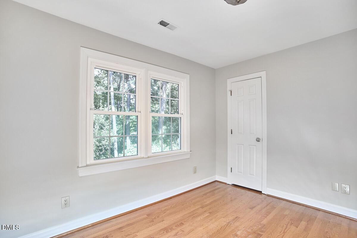 Vacant bedroom with hardwood floors and two windows overlooking leafy trees.