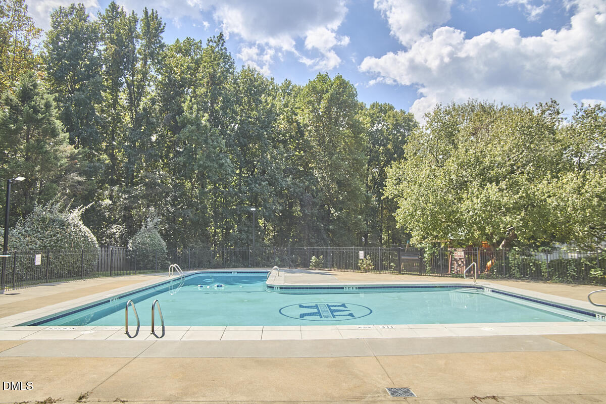 Outdoor swimming pool with a concrete deck, safety fence, and mature trees.