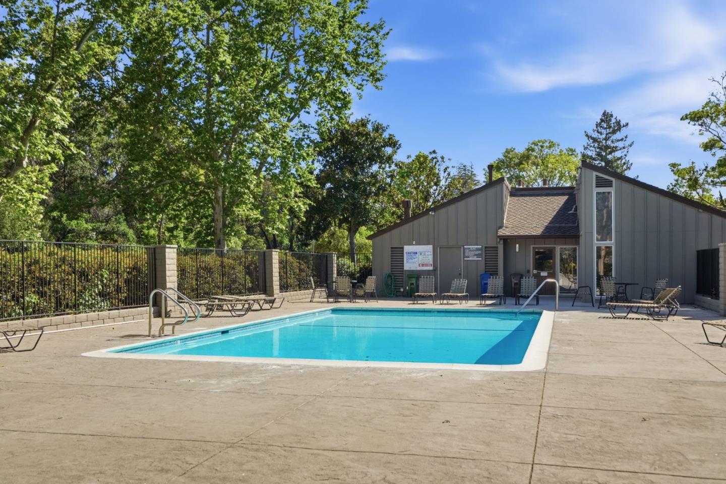Community swimming pool beside a gray pool building, with lounge chairs and mature trees.