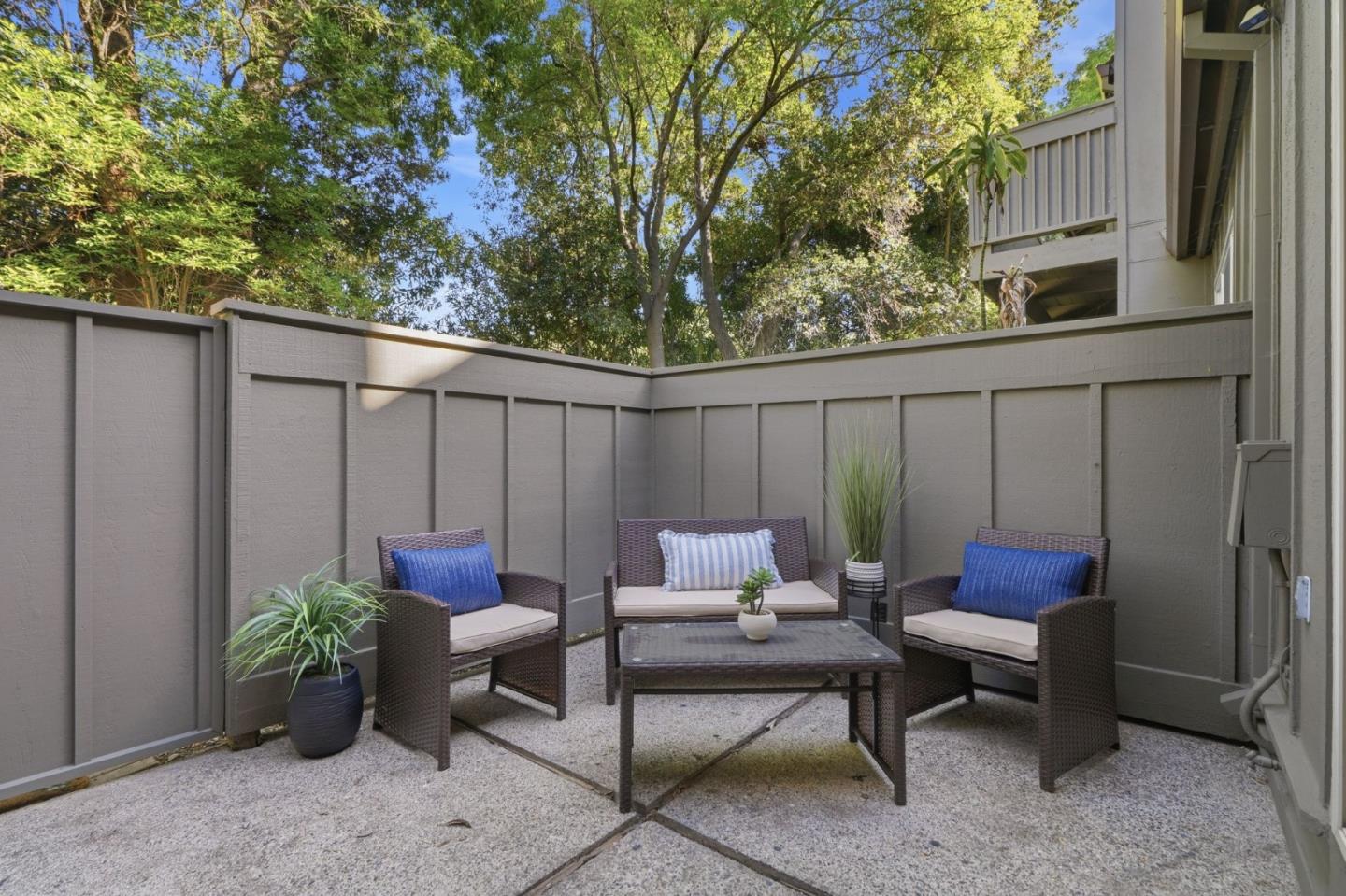 Fenced patio with wicker seating, a glass-top table, and potted plants beneath mature trees.