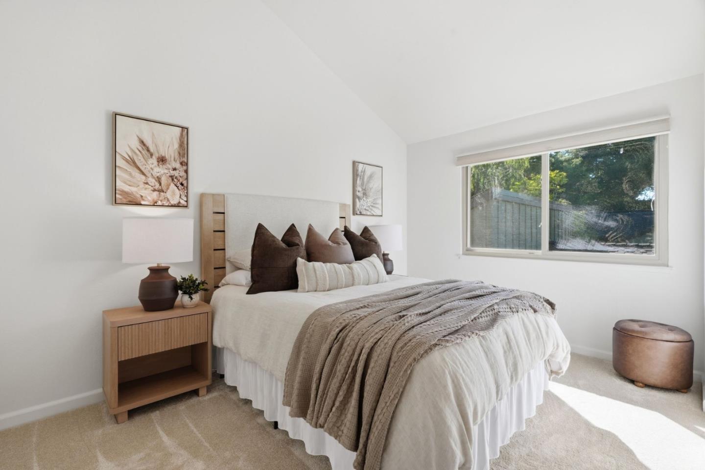 Staged bedroom with a white bed, layered neutral pillows, wooden nightstand, and large window.