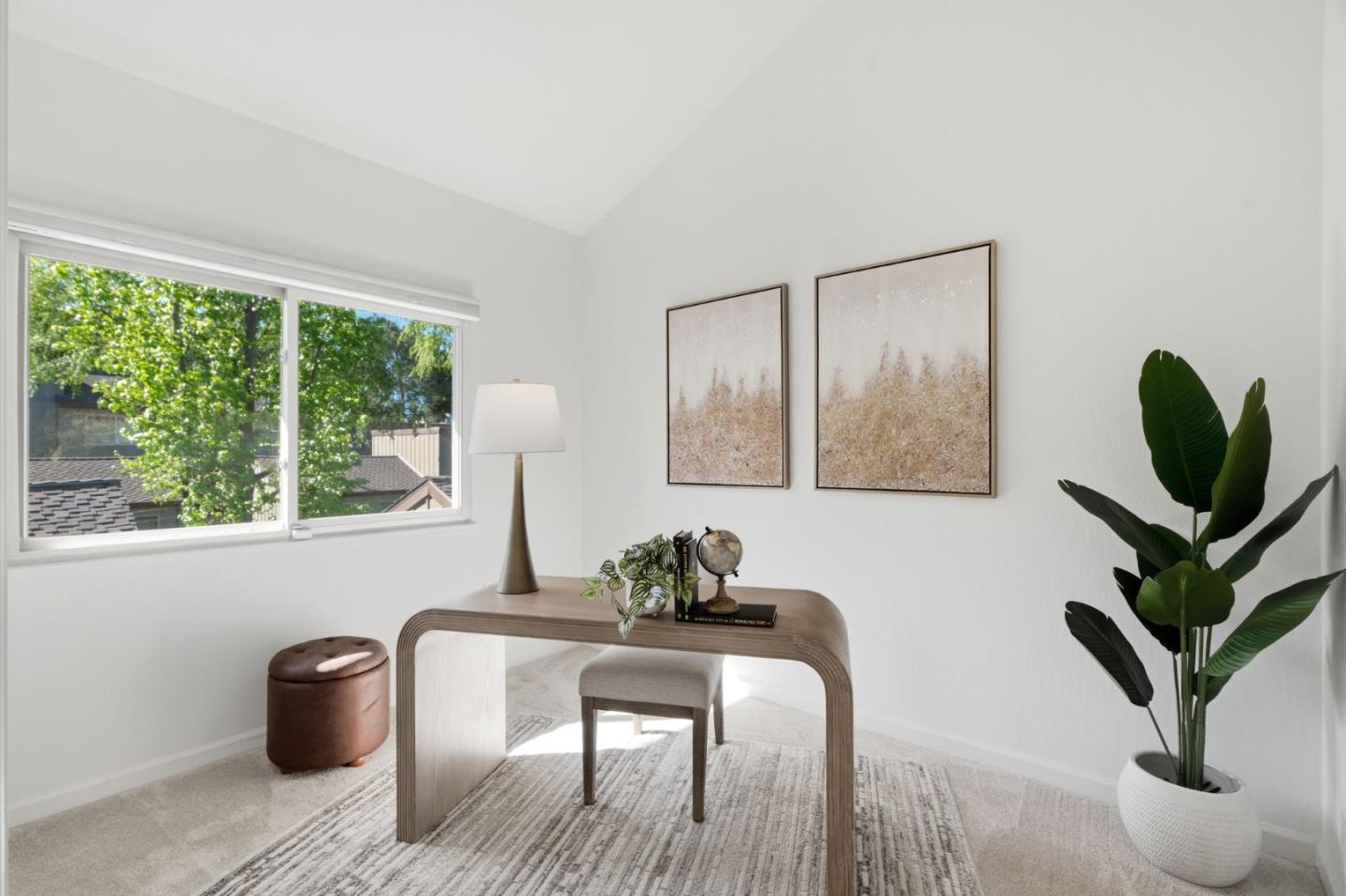 Bright home office with a curved wooden desk, large leafy-window view, framed art, and potted plant.