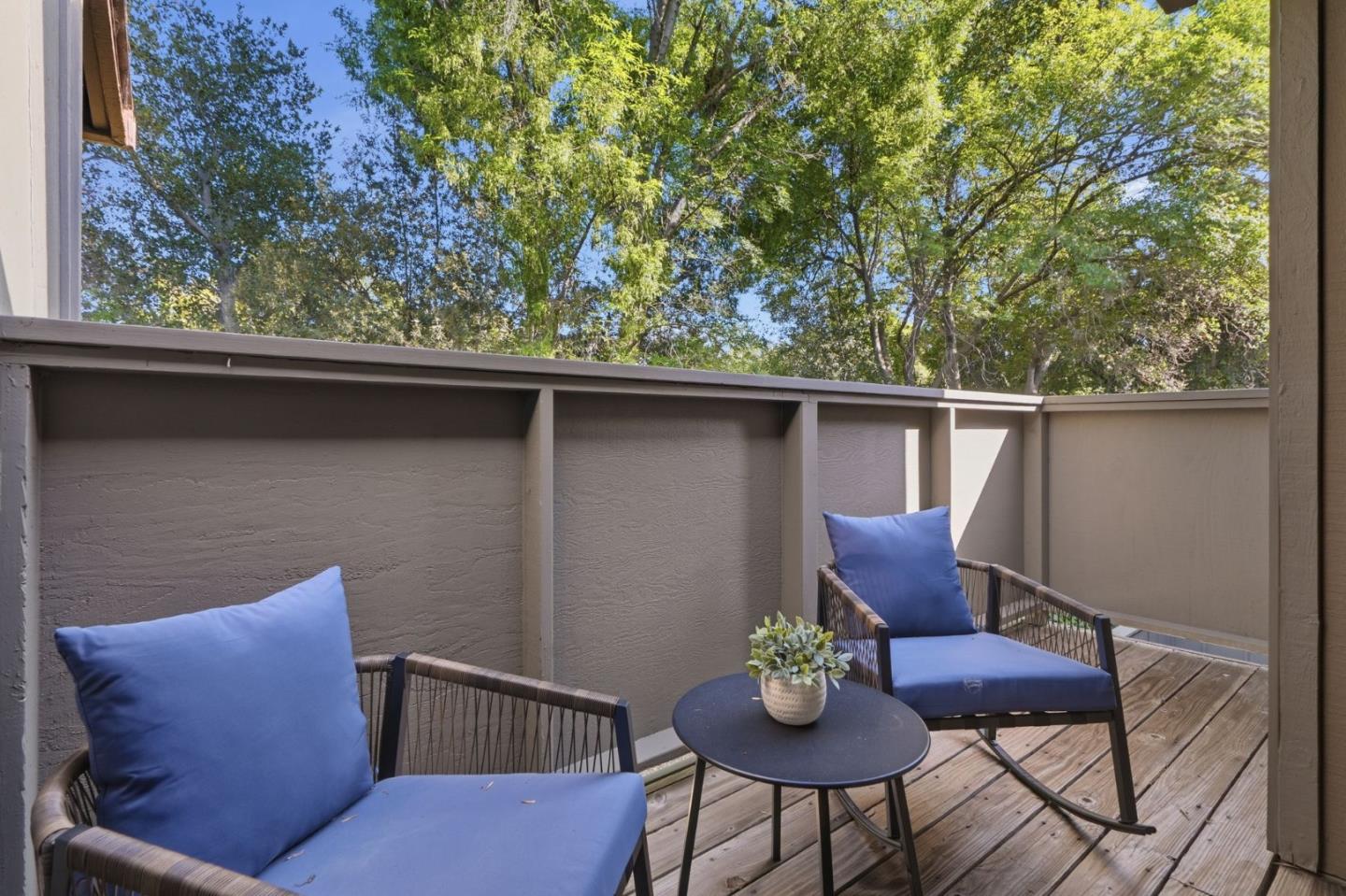 Wood deck with blue-cushioned lounge chairs, a small table, and leafy trees beyond the privacy railing.